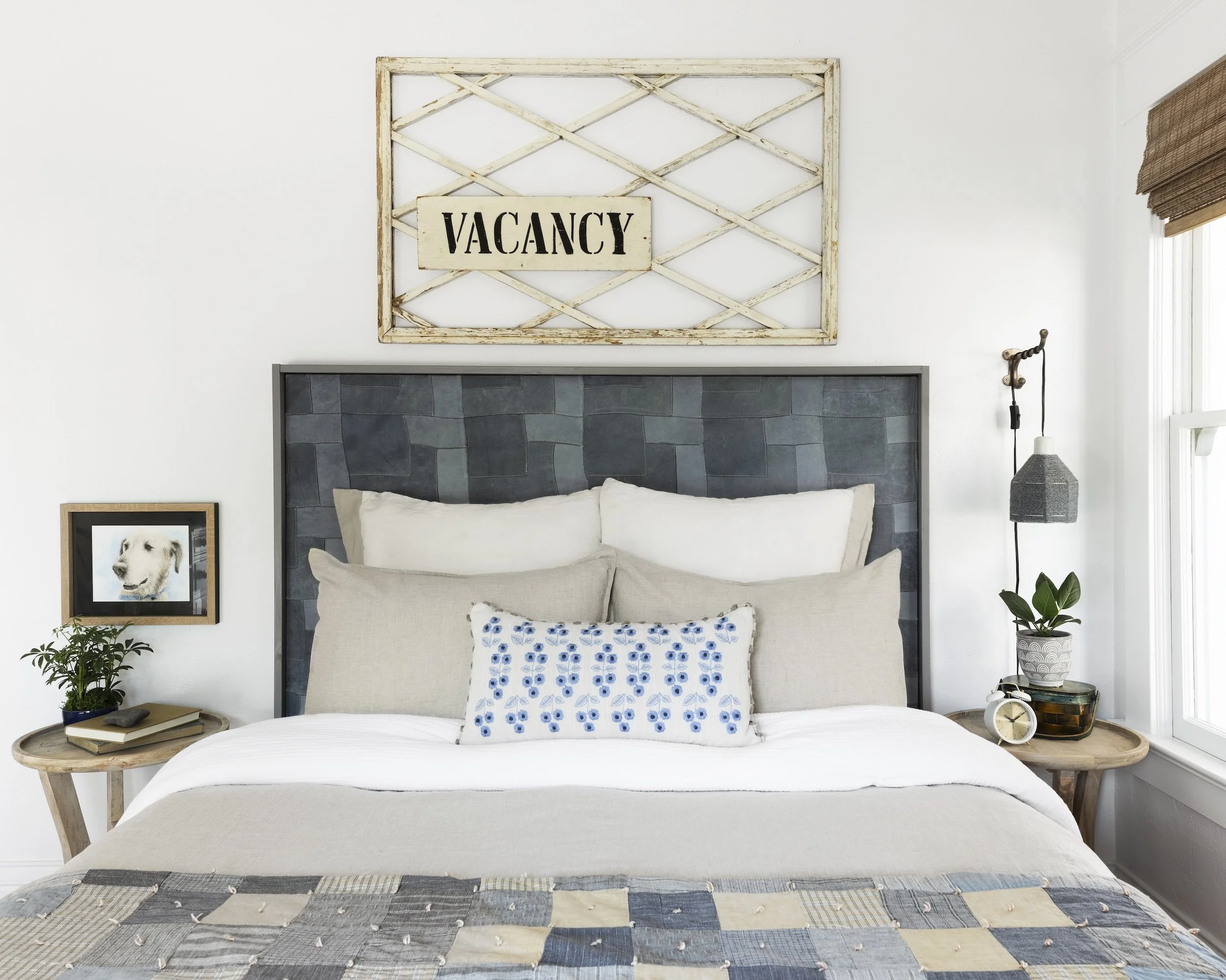 A cozy bedroom with a bed, pillows, and a headboard. Above the bed is a rustic wooden frame with a "Vacancy" sign. On each side of the bed are wooden nightstands with decorative items, including a potted plant, books, a clock, and a picture of a dog.