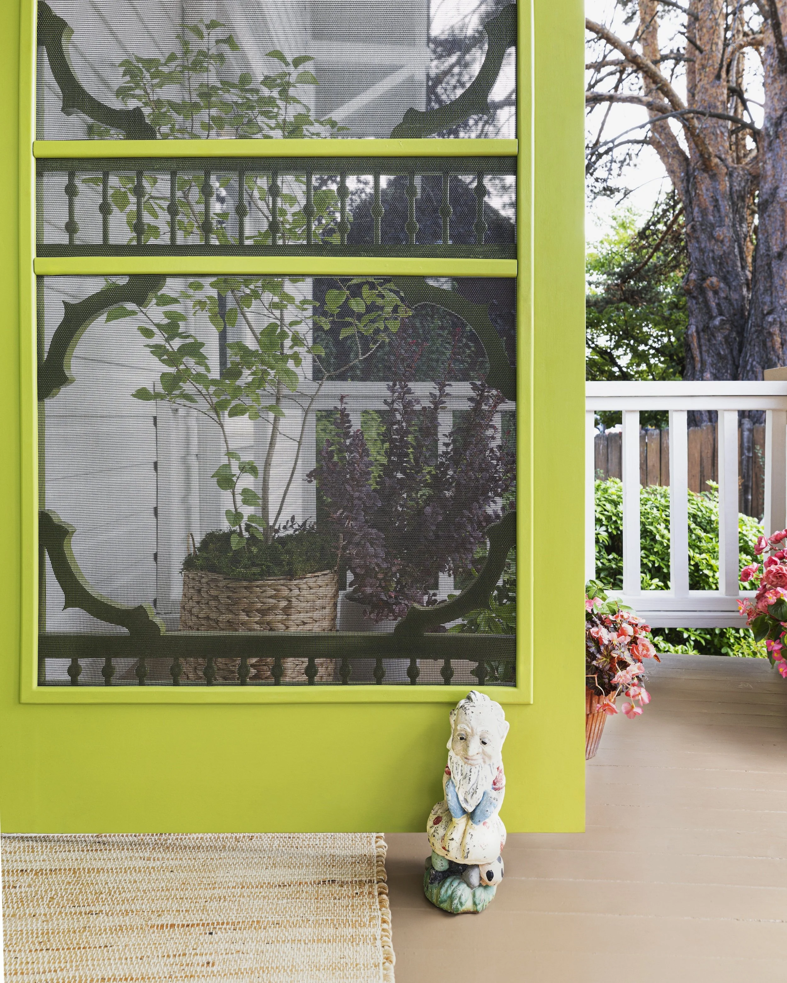 Green screen door with mesh, revealing potted plants and garden outside, with decorative garden gnome figurine on porch with pink flowers nearby.
