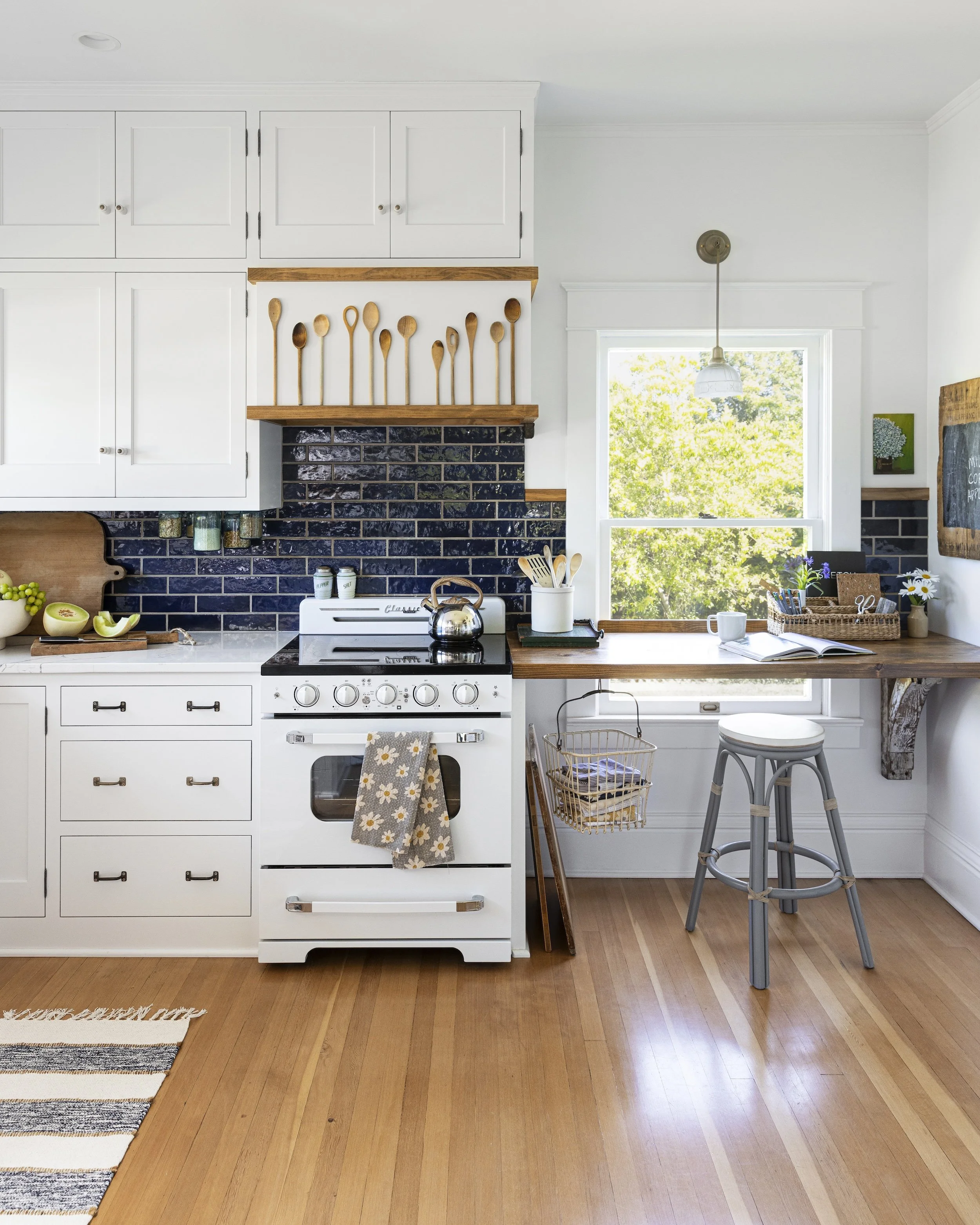 Cozy kitchen with white cabinets, black backsplash, and wooden accents. A vintage stove with a towel, a window showing green trees, and a wooden countertop with various kitchen items.