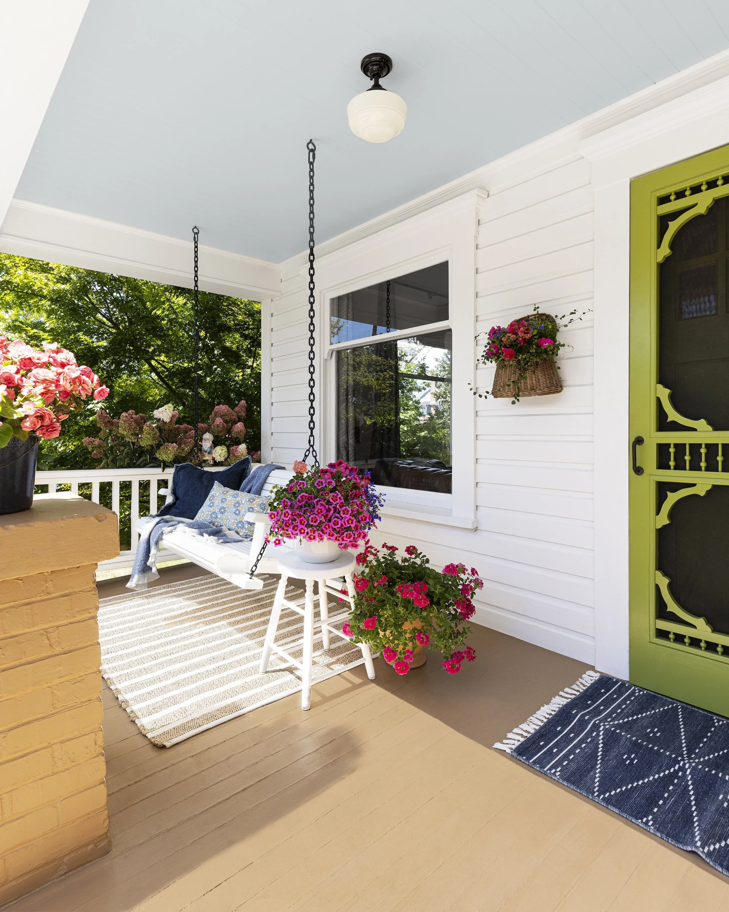 Covered porch with hanging flower basket, white porch swing with cushions, potted pink flowers, and a green door with decorative trim.