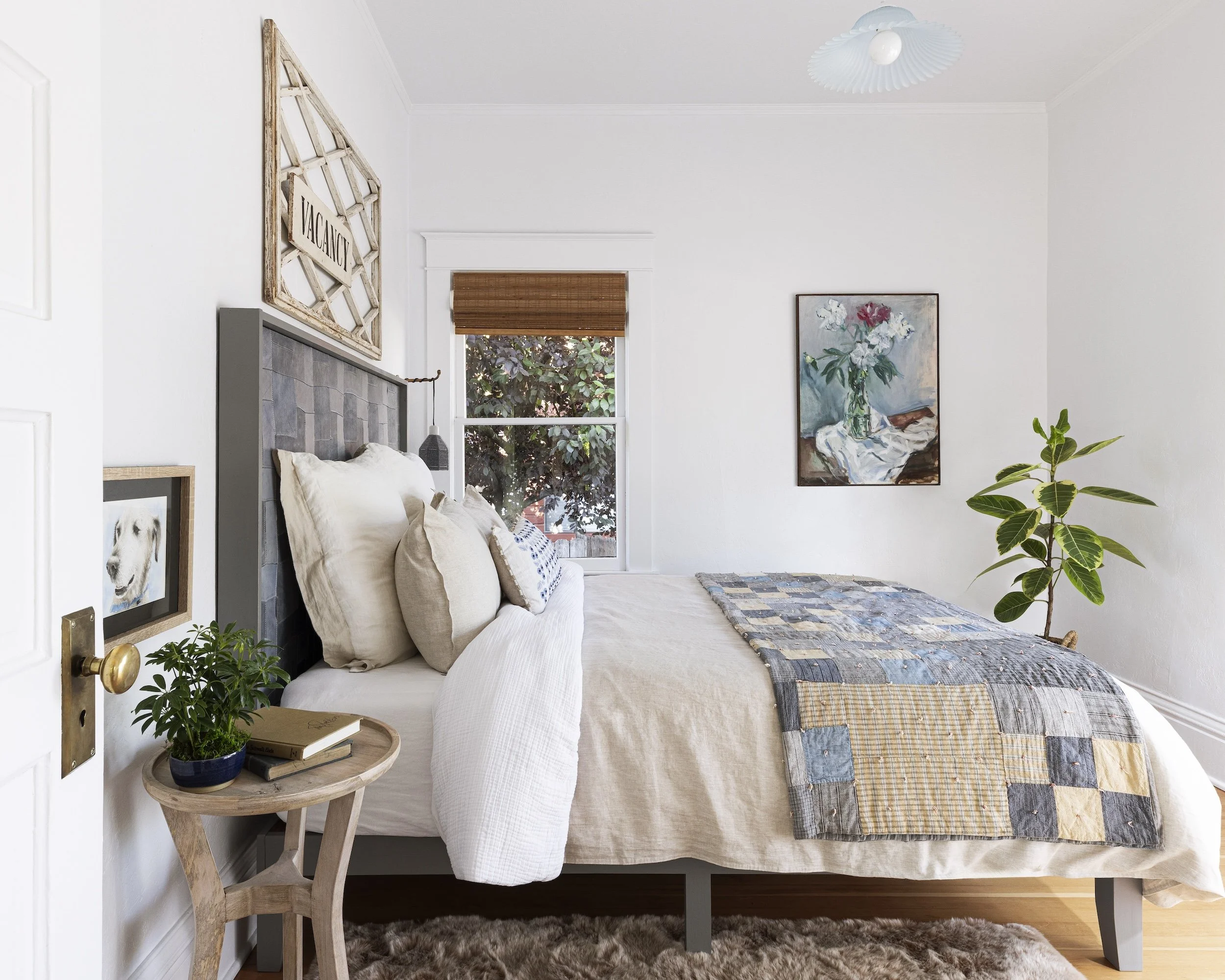 A cozy bedroom with a window, white walls, and a bed with beige and white bedding and throw pillows. There's a small wooden side table with a potted plant and books, a framed dog portrait on the wall, a floral painting on the wall, and a tall leafy p
