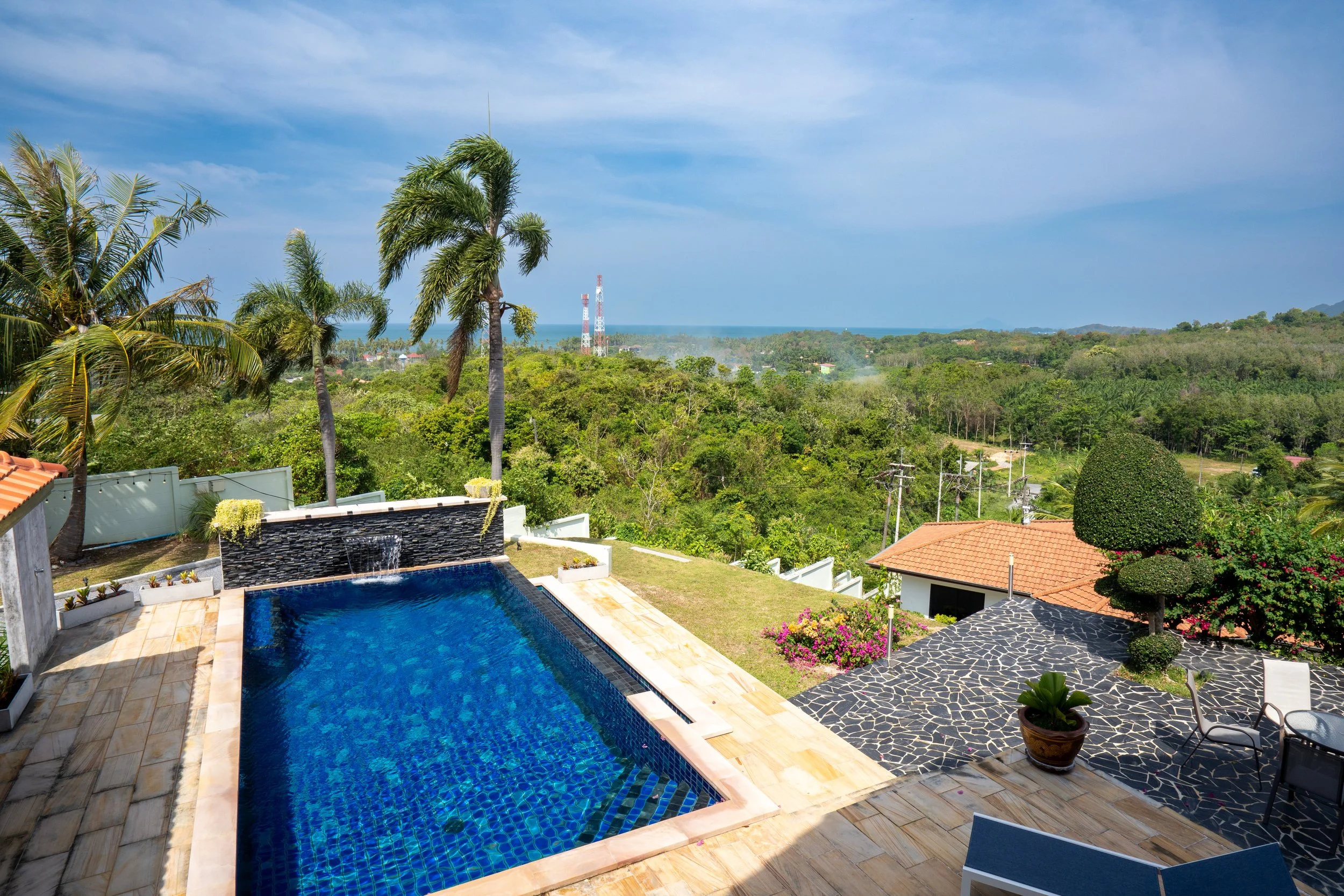 View of a swimming pool surrounded by a patio with lounge chairs, potted plants, and well-manicured trees, overlooking a lush green landscape with palm trees, distant ocean, and power lines under a blue sky.