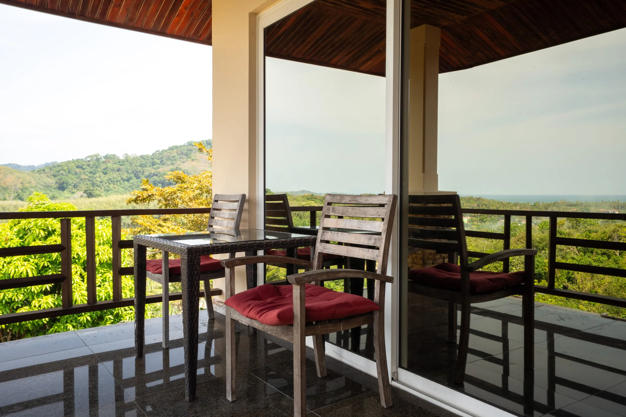 Balcony with wooden chairs and a glass-top table overlooking green trees and distant hills.