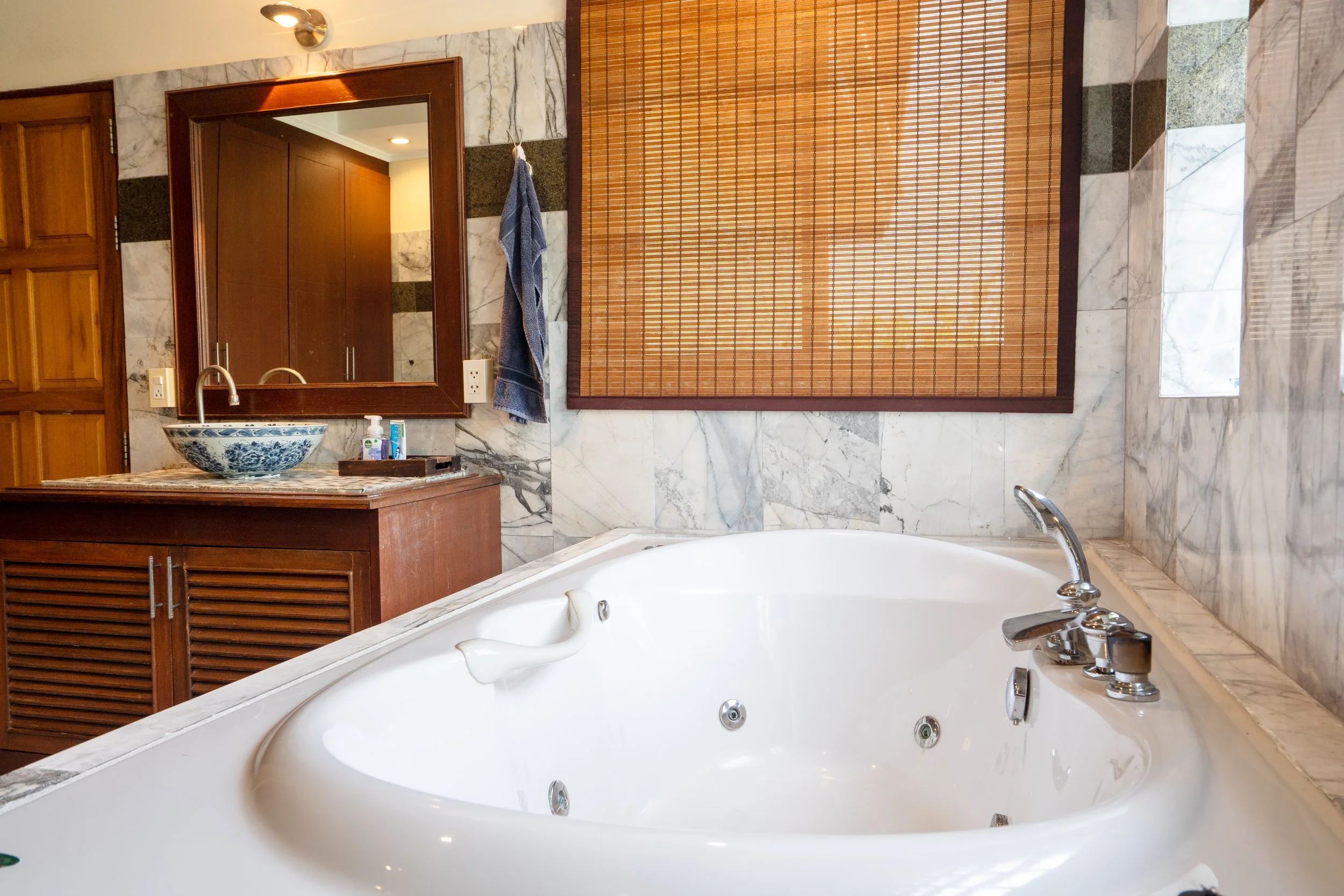 A bathroom featuring a large whirlpool bathtub with chrome fixtures, surrounded by marble tiles. The room has a wooden vanity with a decorative bowl sink, a mirror, a towel hanging near the mirror, and a window with bamboo blinds.