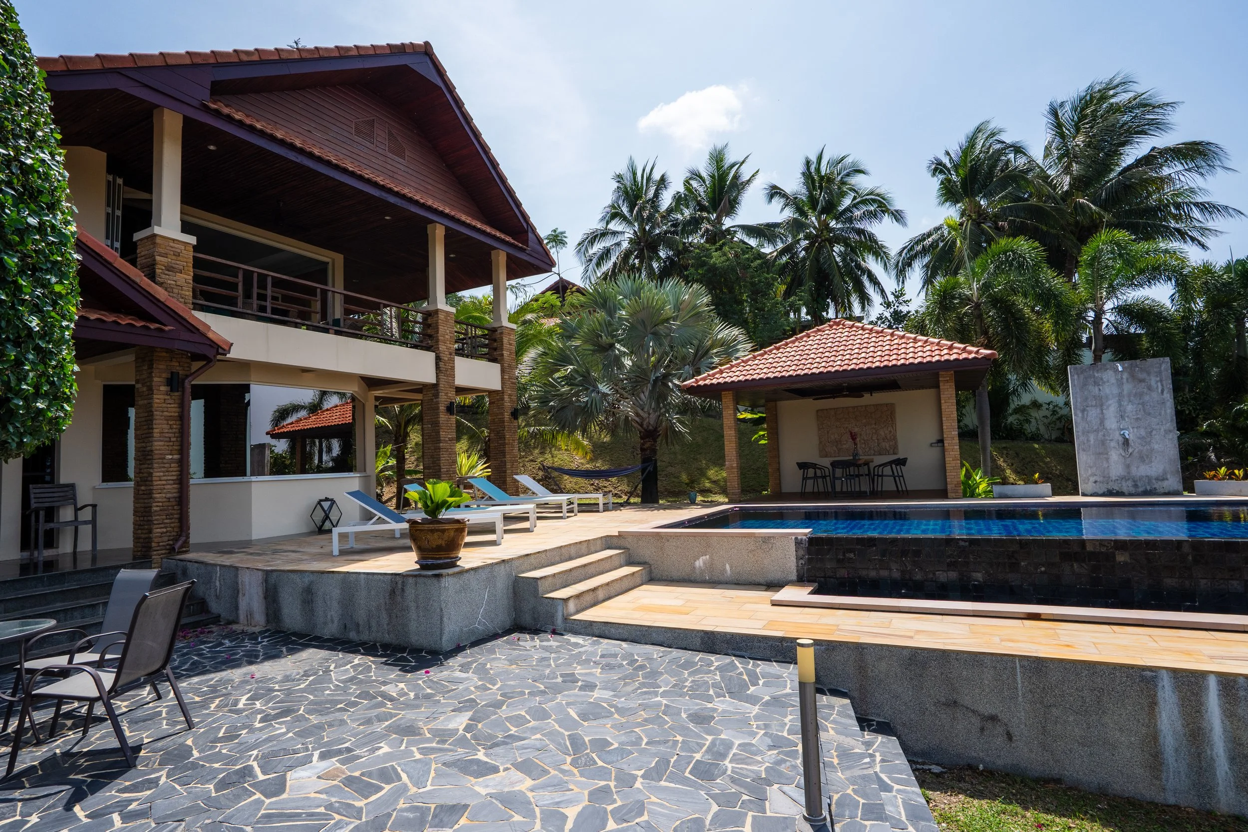 Luxury villa backyard with a swimming pool, sun loungers, a shaded seating area, lush palm trees, and tropical plants under a clear sky.