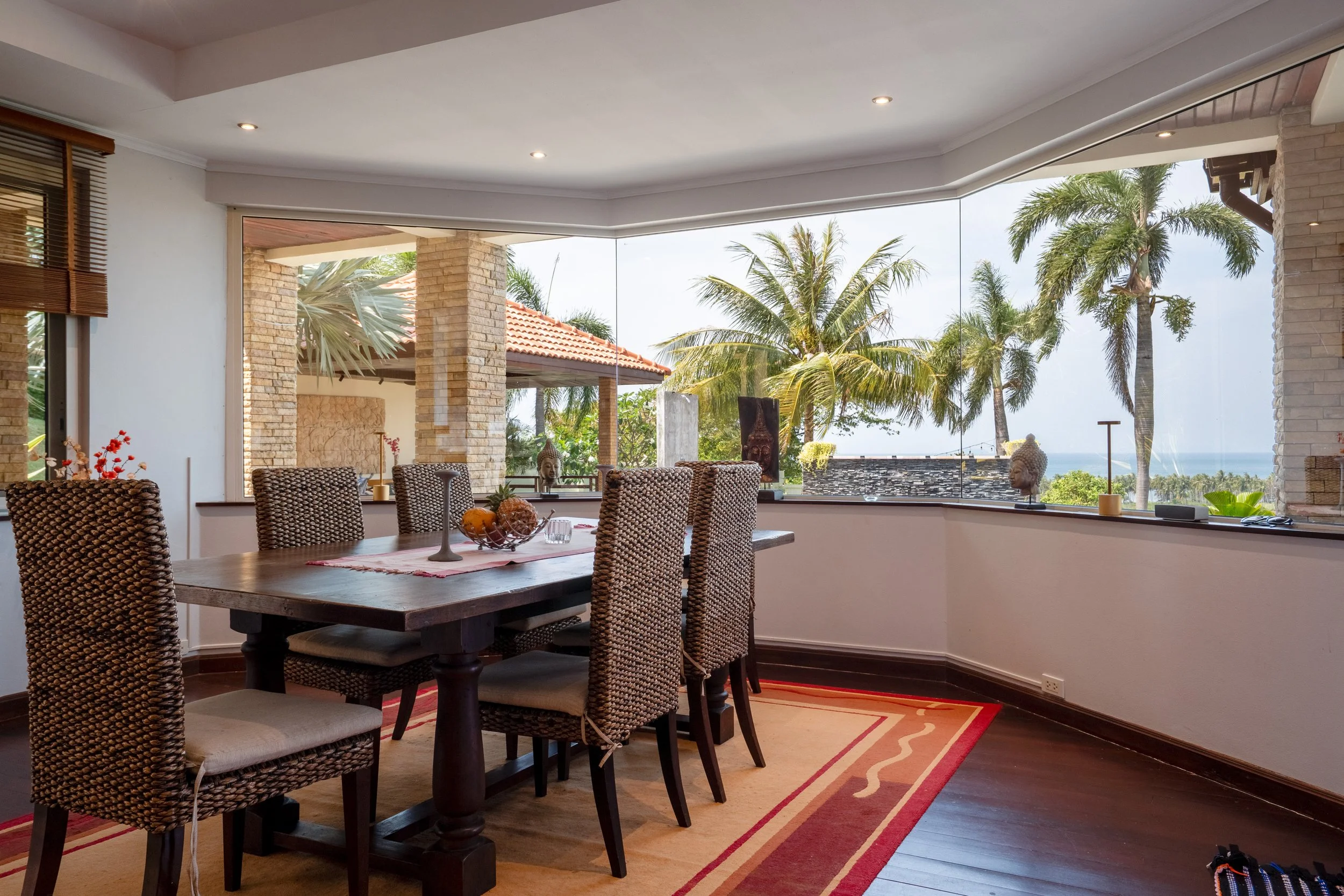 A dining room with a wooden table, six woven chairs, and a view of palm trees and the ocean outside through large windows.