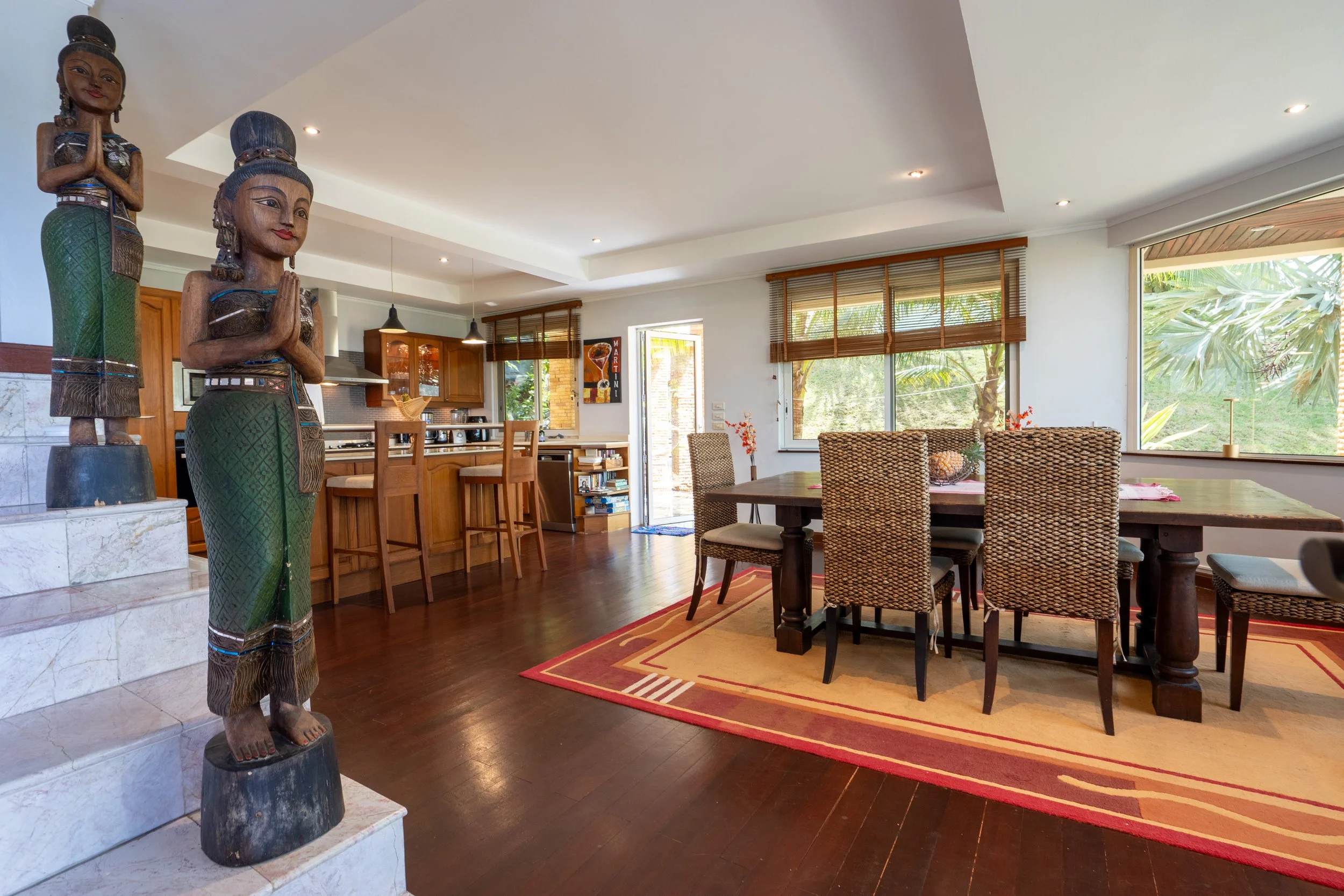 Living room with wooden accents, large windows, wicker chairs around a wooden dining table, colorful patterned area rug, and traditional carved wooden statues near stairs.