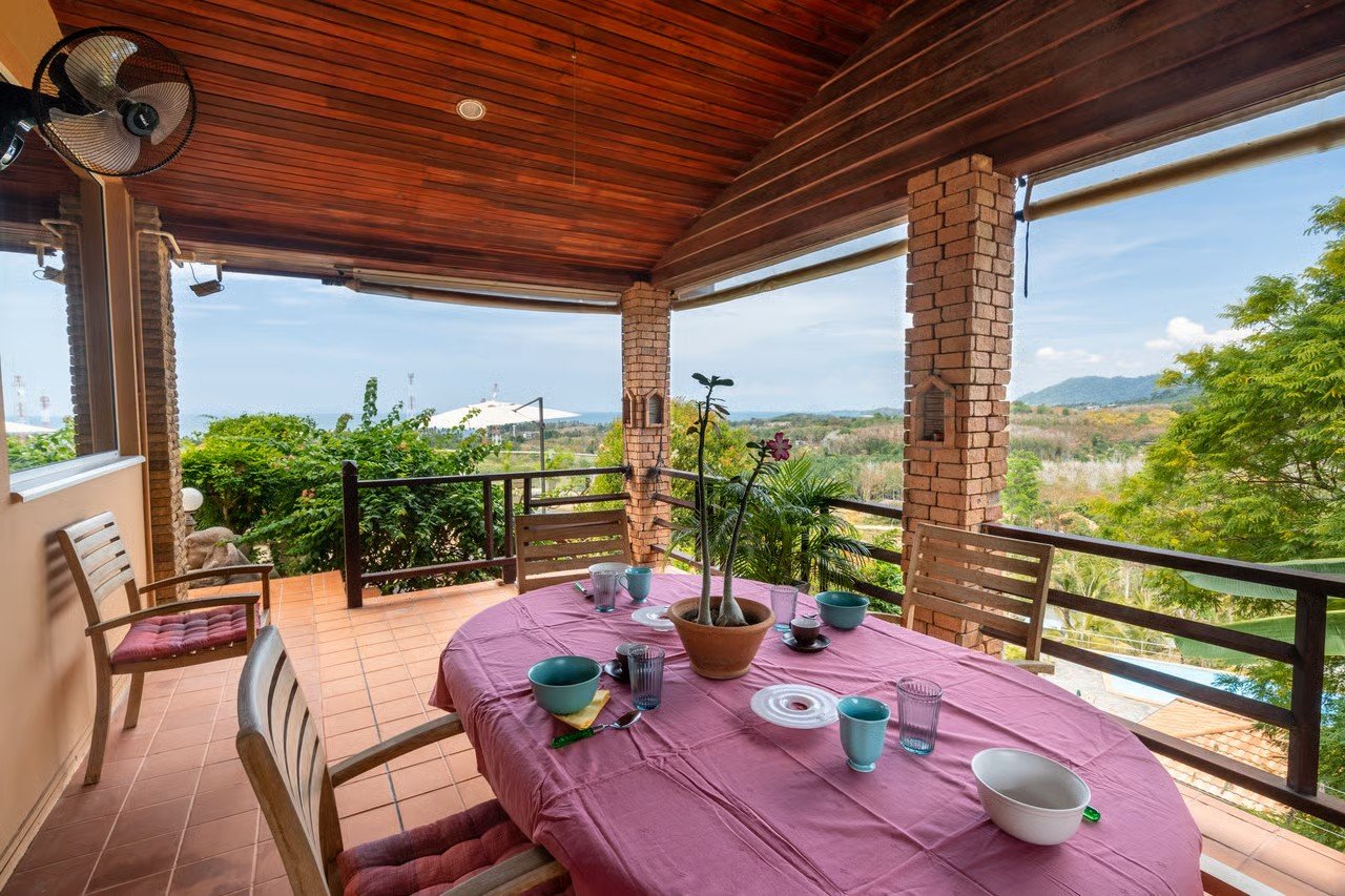 Outdoor dining area with a pink tablecloth, bowls, glasses, and a potted plant, overlooking trees and hills in the distance.