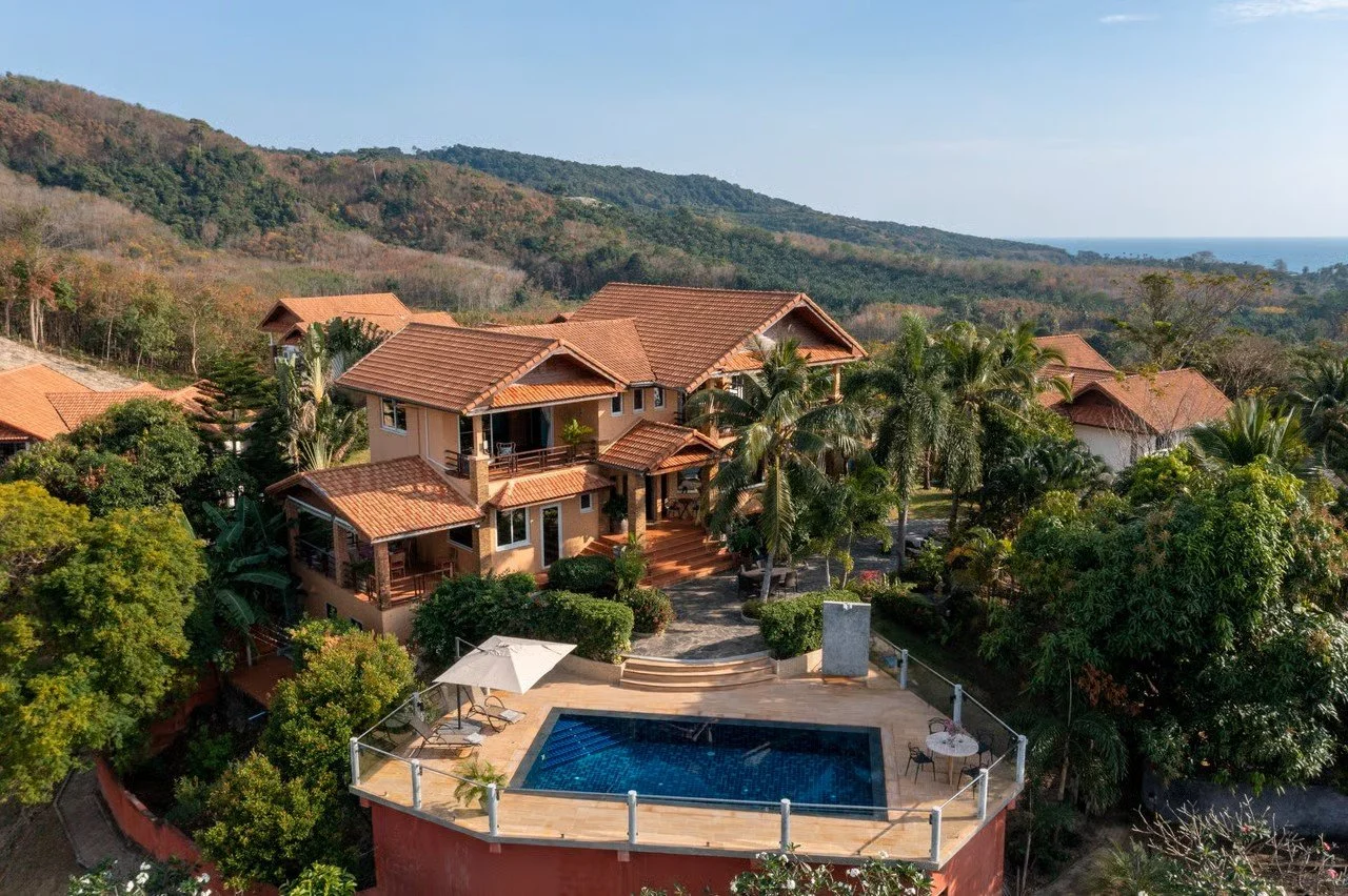 A large house with a red tile roof, surrounded by palm trees, lush greenery, and a pool with an umbrella and lounge chairs in the foreground, with hills and ocean in the background.