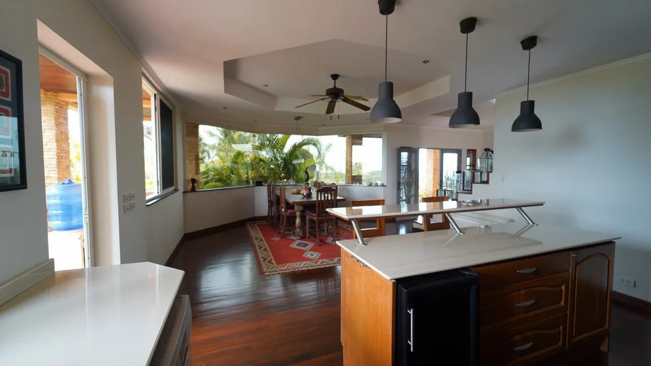 Open-plan kitchen and dining area with large windows showing outdoor greenery, dark wood floors, a kitchen island with pendant lights above, and a dining table with chairs.