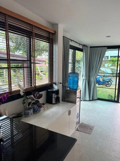 Interior view of a kitchen and living area with large windows, a water jug, a coffee machine, potted plants, and sliding glass doors leading to a backyard with a motorcycle and greenery.