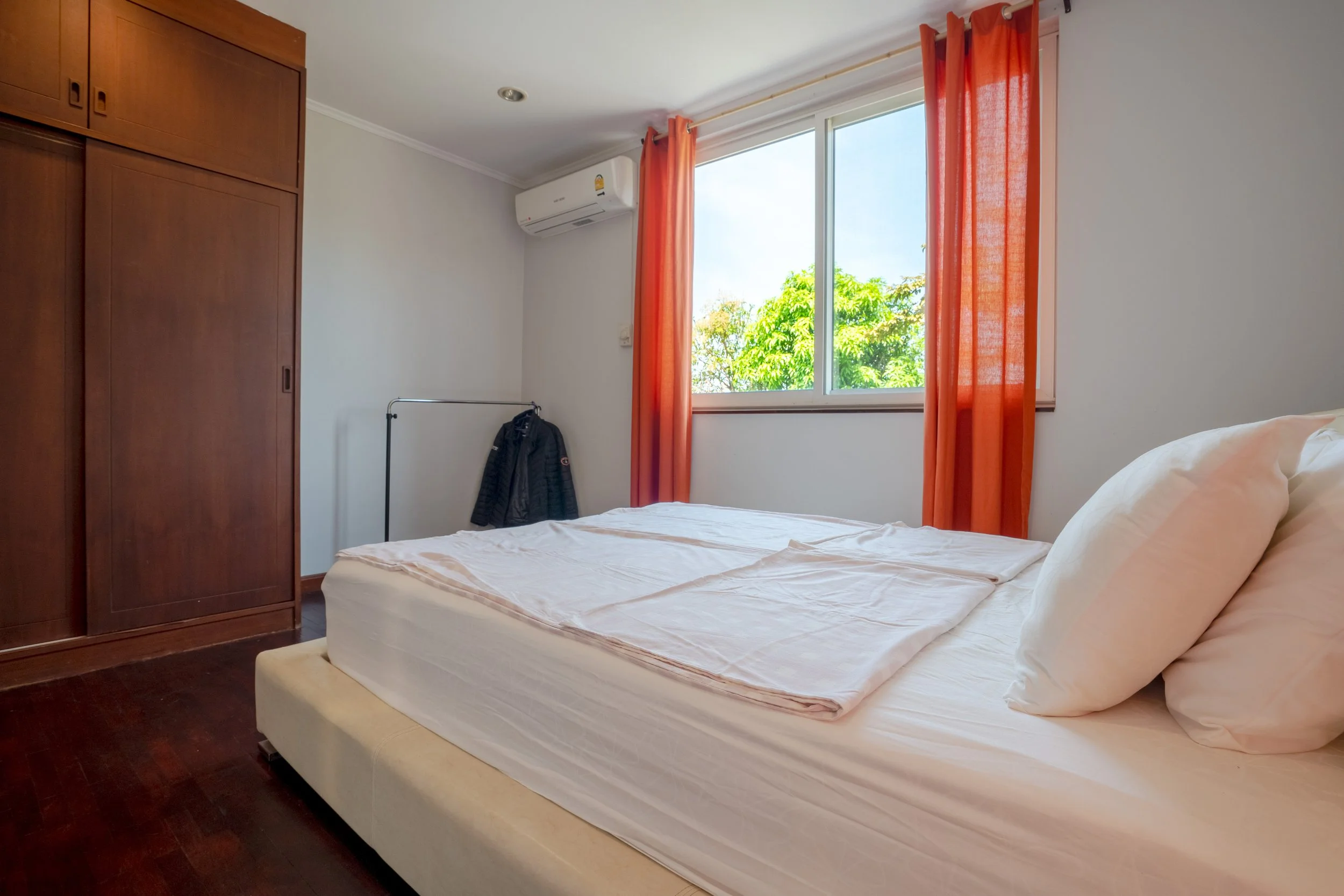 A simple bedroom with a bed, orange curtains, a wooden wardrobe, and an window showing greenery outside.