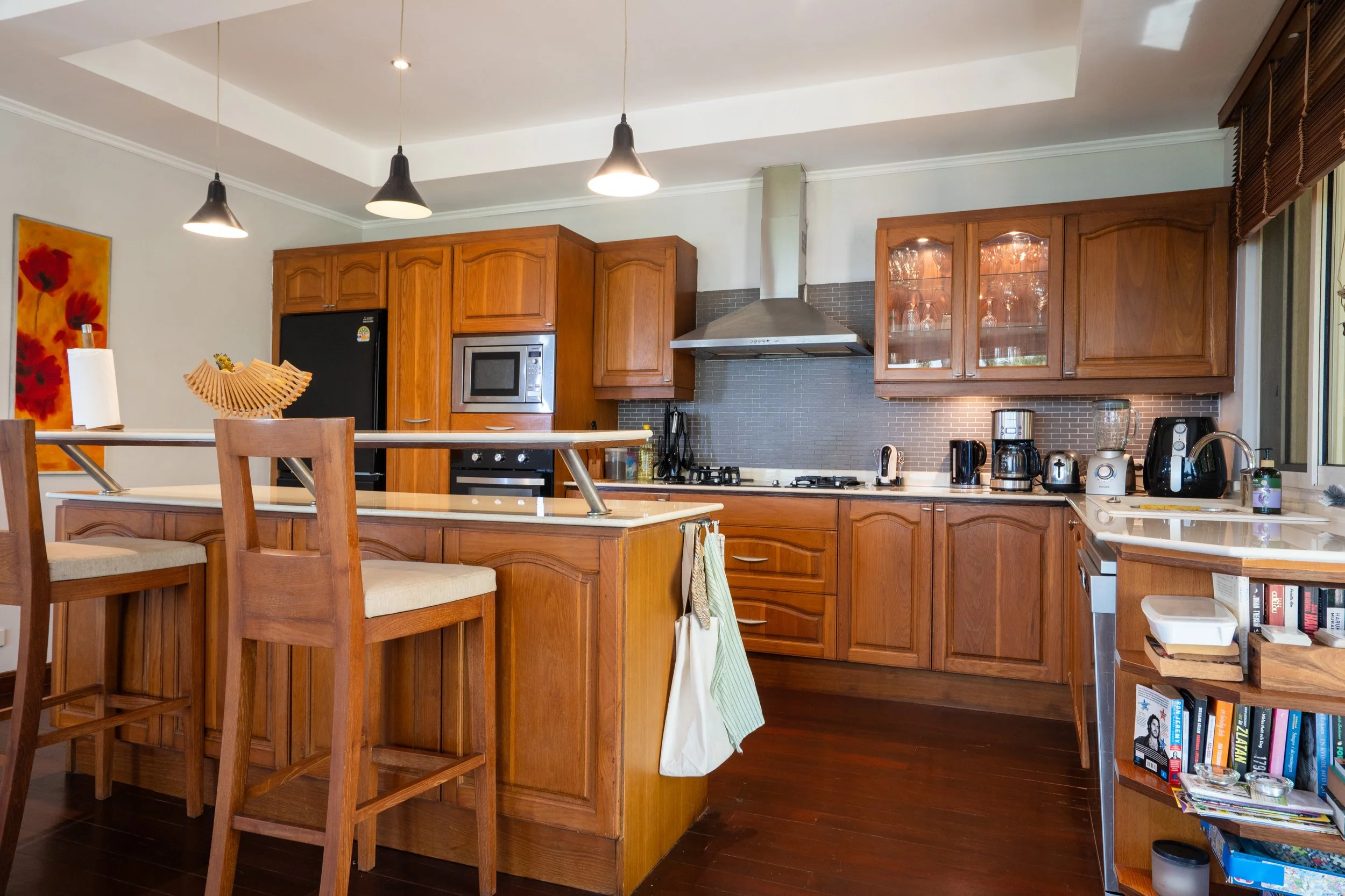 Kitchen with wooden cabinets, a black refrigerator, microwave, and various appliances on the countertop. There is an island with bar stools and a wall with books and kitchen items.