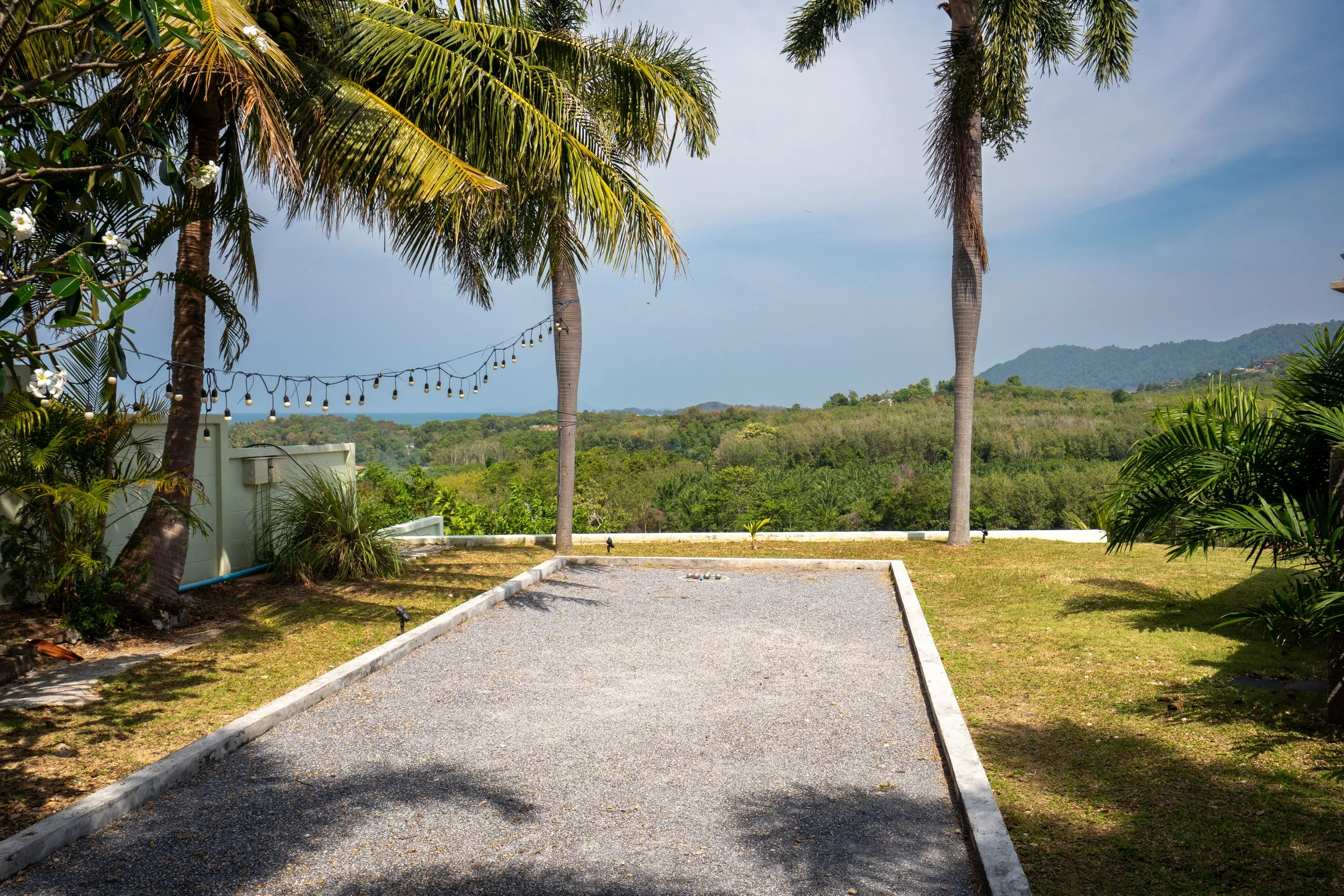 A backyard with a gravel bocce court, surrounded by tropical plants and trees, overlooking a lush landscape with hills and a partly cloudy sky.