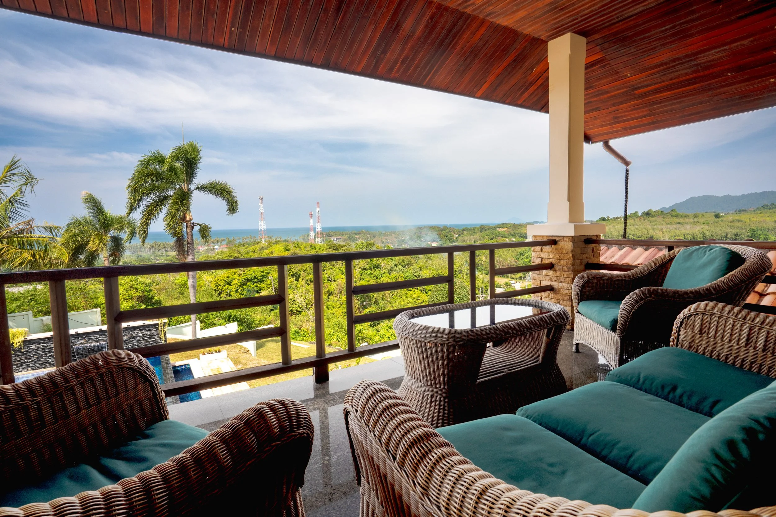 View from a balcony with wicker chairs and teal cushions overlooking lush green trees, palm trees, and distant hills under a partly cloudy sky.