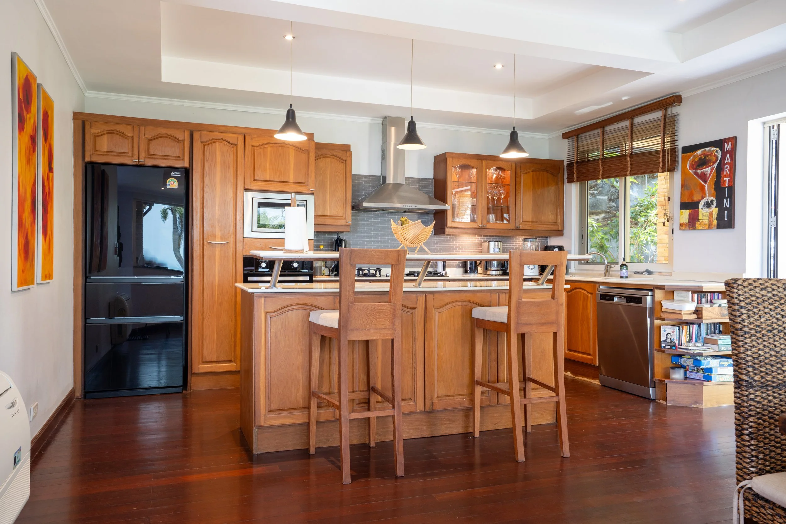 Kitchen with wooden cabinets, an island with barstools, black refrigerator, and a window with blinds, decorated with colorful paintings and a bookshelf.
