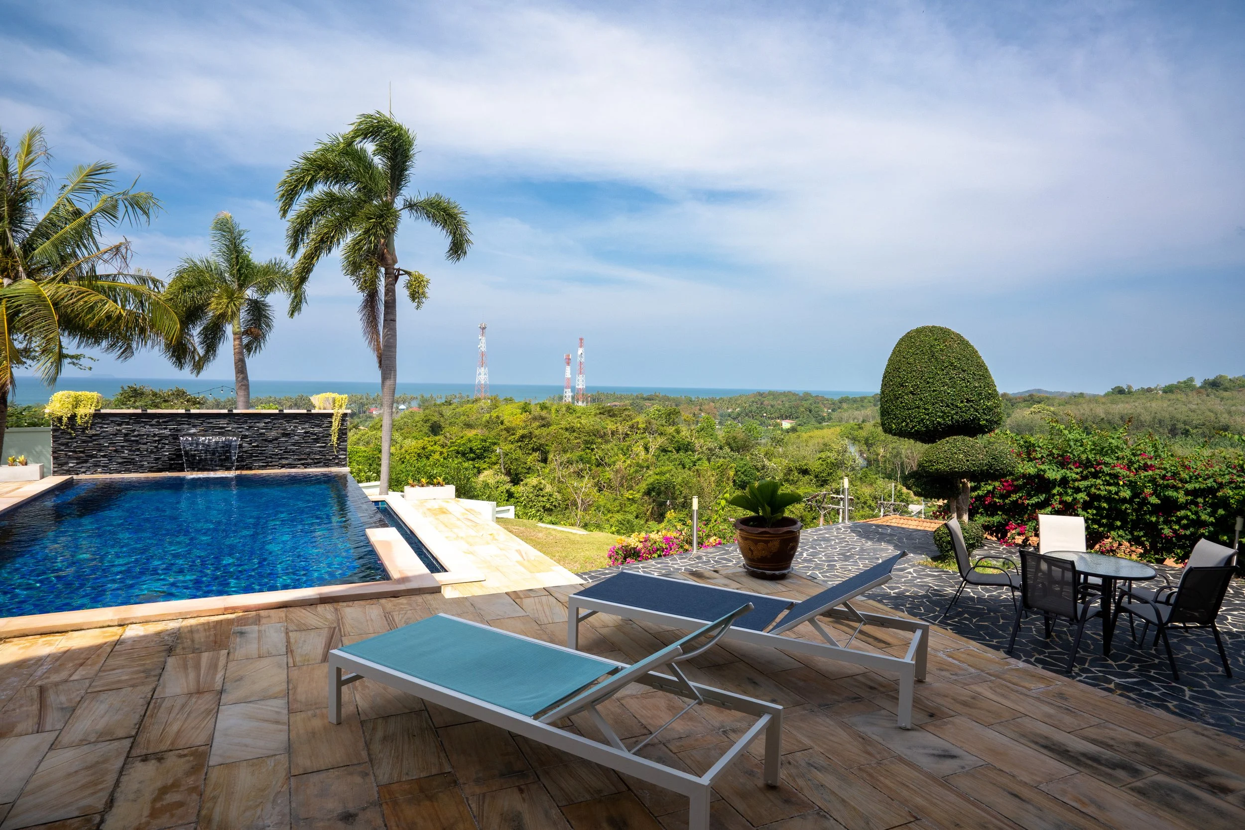Outdoor pool area with lounge chairs, a table with chairs, potted plants, palm trees, and a view of trees and the ocean in the distance.