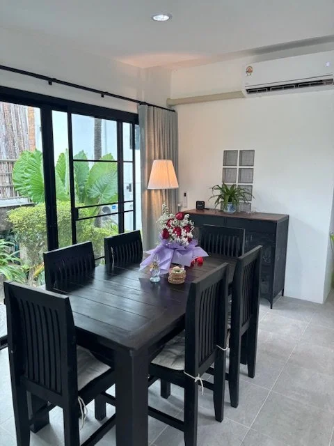 Dining room with black table and chairs, bouquet of flowers, sliding glass door, curtains, sideboard with plant and decorative items, air conditioning unit, floor lamp, and greenery outside.