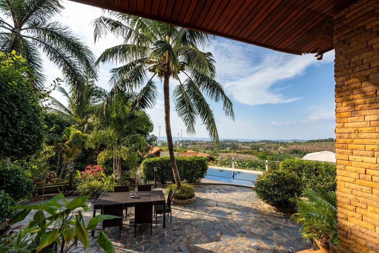 Outdoor patio with a table and chairs, surrounded by lush tropical plants and palm trees, overlooking a scenic landscape with a pool and distant hills.