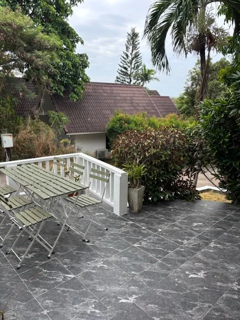 Outdoor patio with a white railing, a glass-top table, and four chairs, surrounded by greenery and trees, with a house and cloudy sky in the background.