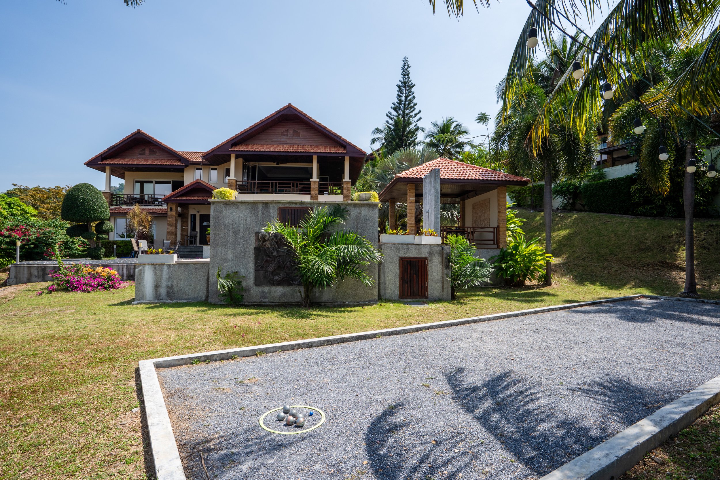 A large tropical house with multiple levels, surrounded by lush green trees and plants, featuring a gravel bocce court with metal balls in the foreground.