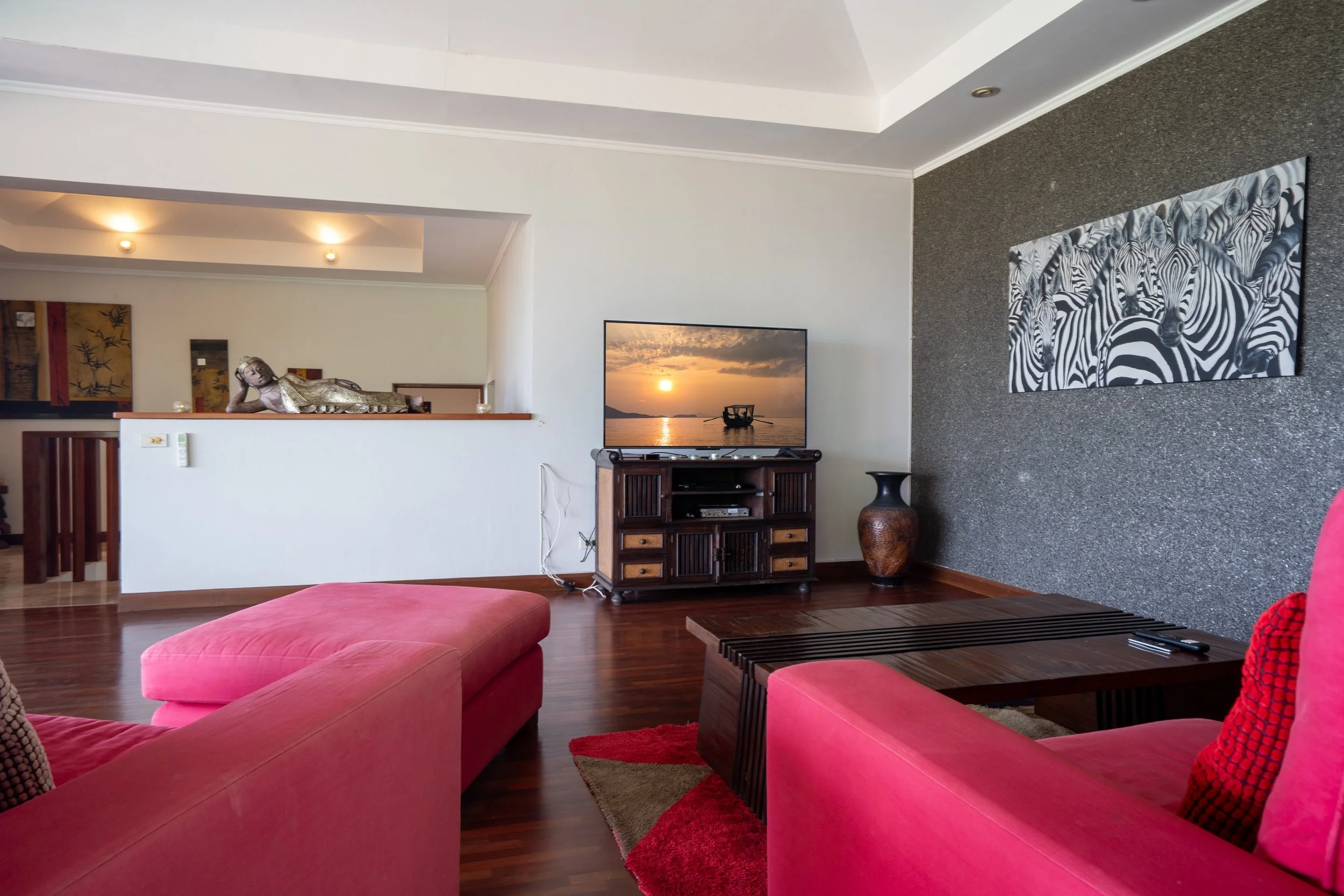 Living room with pink sofas, a flat-screen TV on a wooden stand, a large vase, and a zebra-themed wall art, with a partial view of an open space behind.