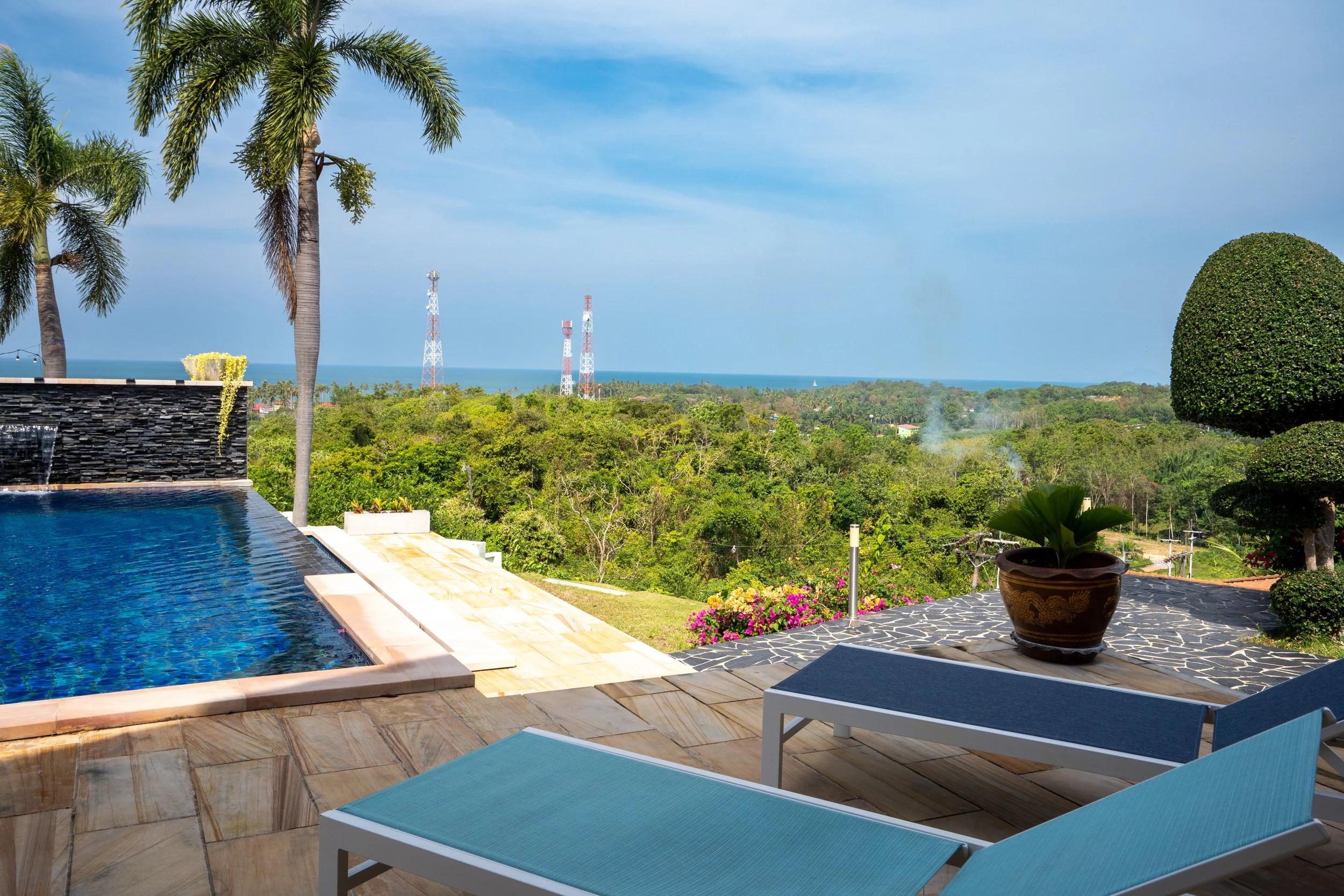 Poolside view with lounge chairs, potted plant, trees, and a lush green landscape with the ocean and blue sky in the background.