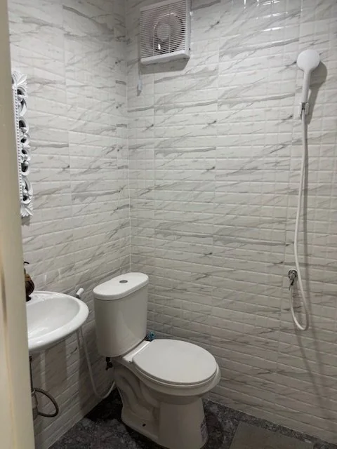 Bathroom with white brick-style tiled walls, a toilet, a small sink, and a handheld shower head with a hose. There is a small exhaust fan on the wall.