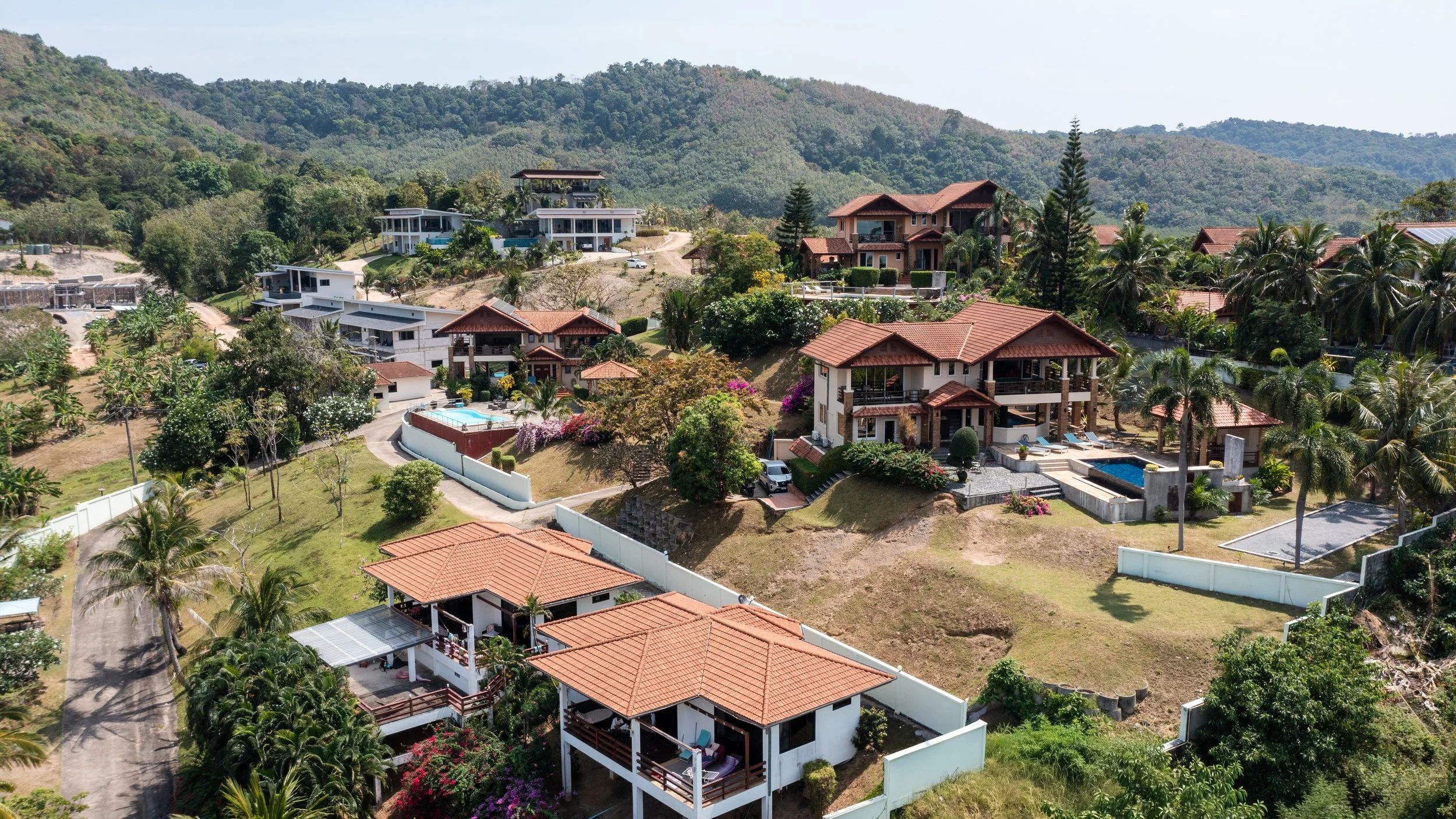 Aerial view of a hillside neighborhood with multiple modern houses, lush greenery, palm trees, and a mountain backdrop.