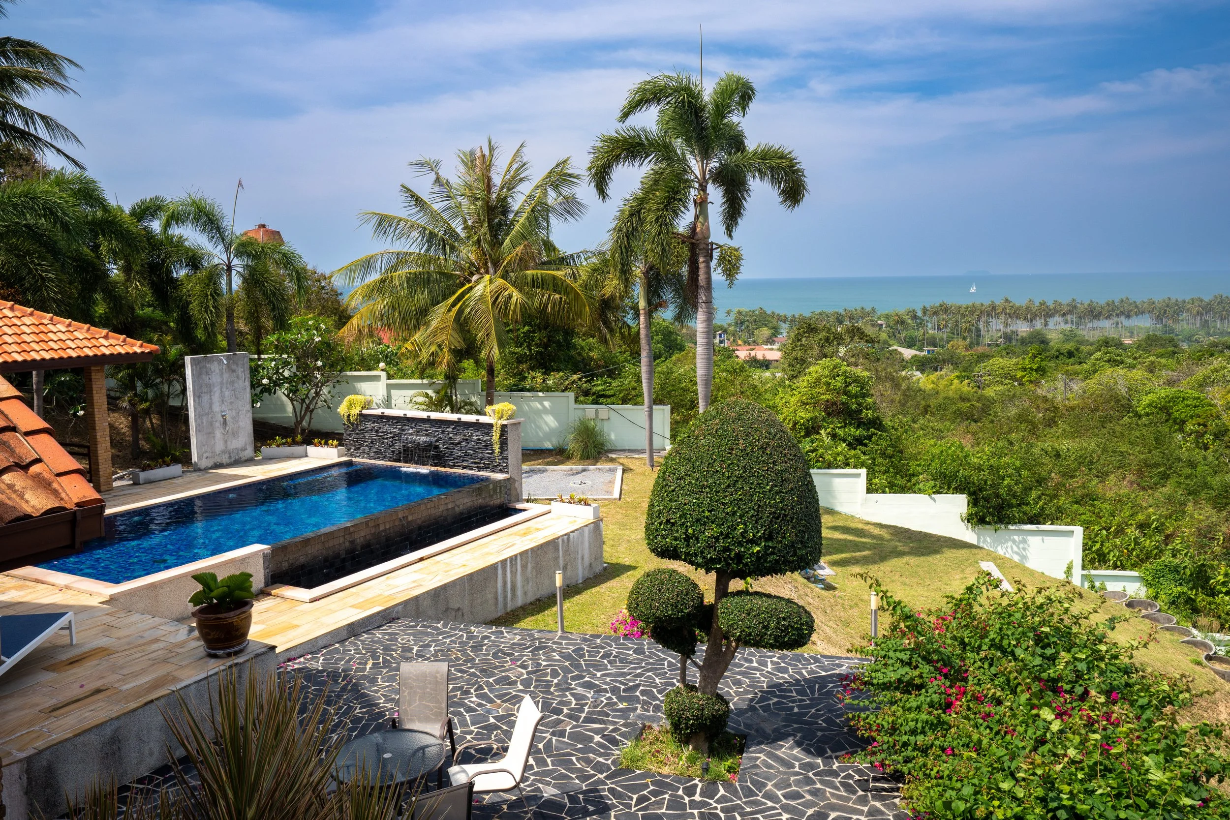 A backyard with a swimming pool, palm trees, and a view of the ocean in the distance.