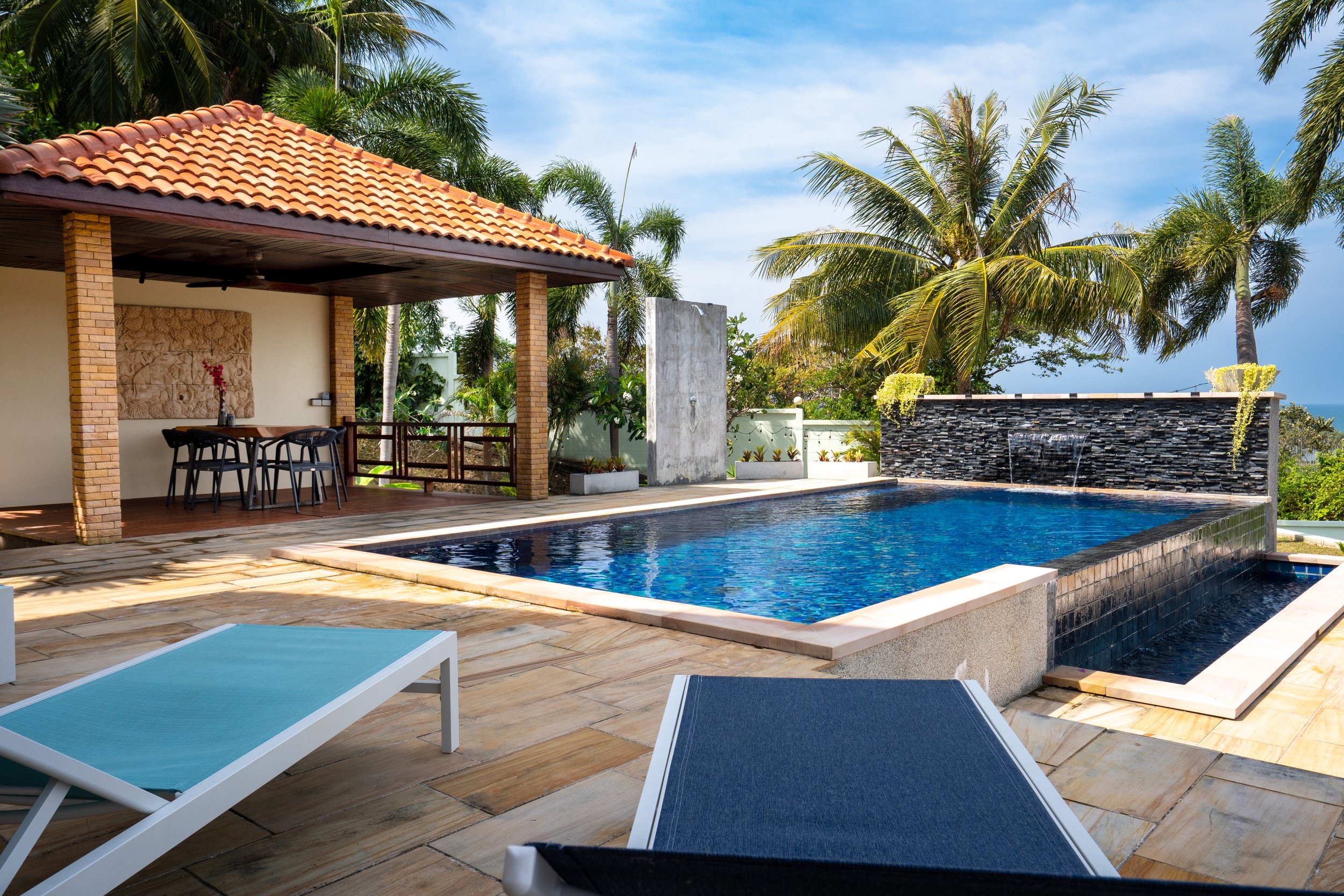 A luxurious outdoor swimming pool area with lounge chairs, a covered seating area with a dining table and chairs, and lush palm trees in the background under a partly cloudy sky.