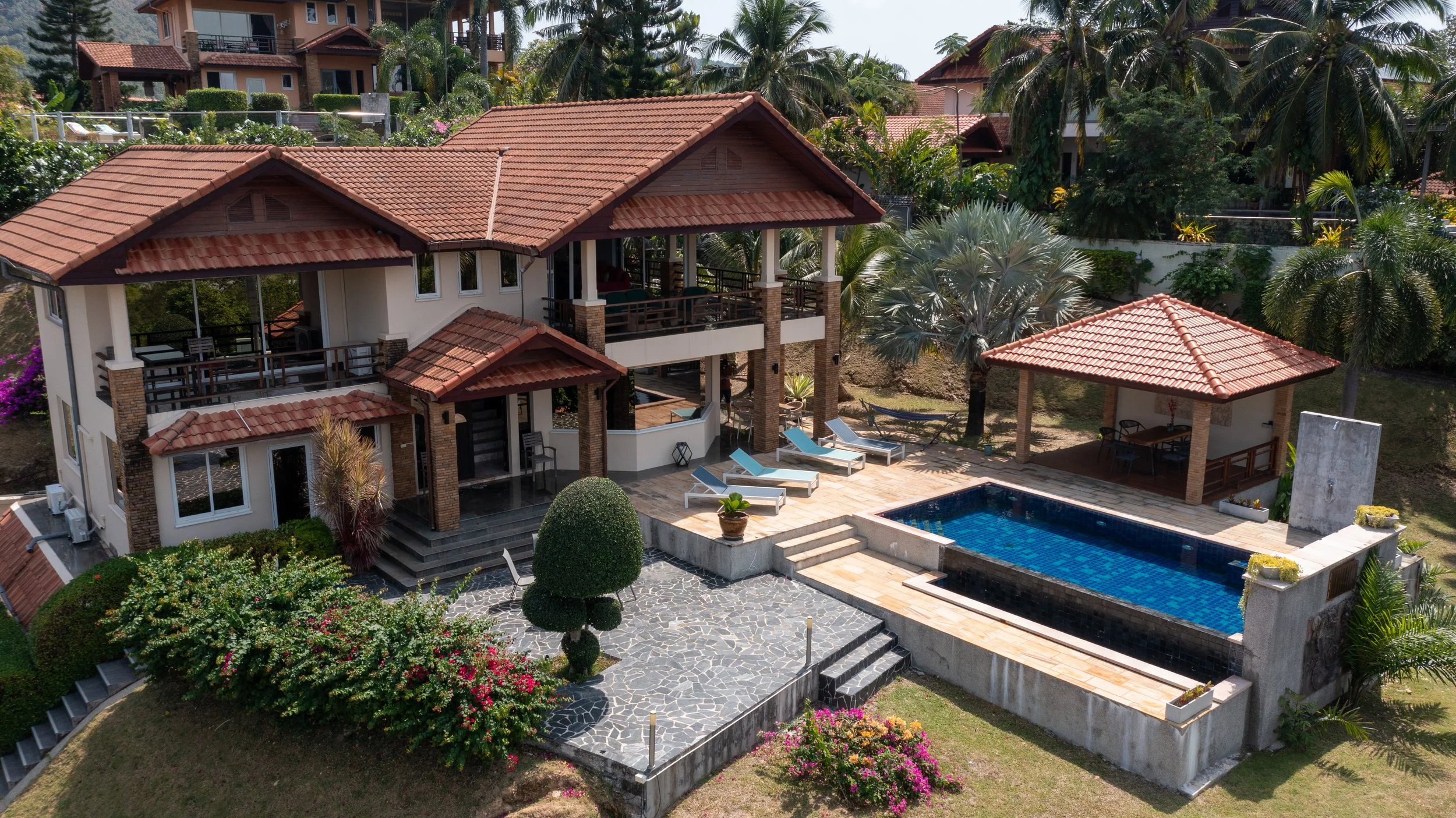 A large house with multiple levels, red tile roofs, and a swimming pool with lounge chairs, surrounded by lush greenery and palm trees.