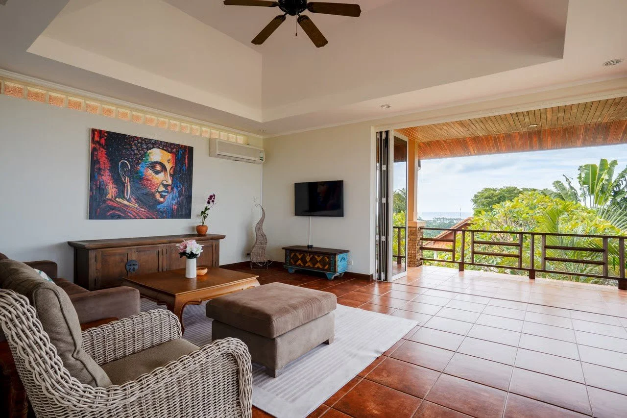 Living room with wicker and fabric furniture, a wooden coffee table, wall art of a colorful Buddha, a TV, and an open balcony with a view of green trees and blue sky.
