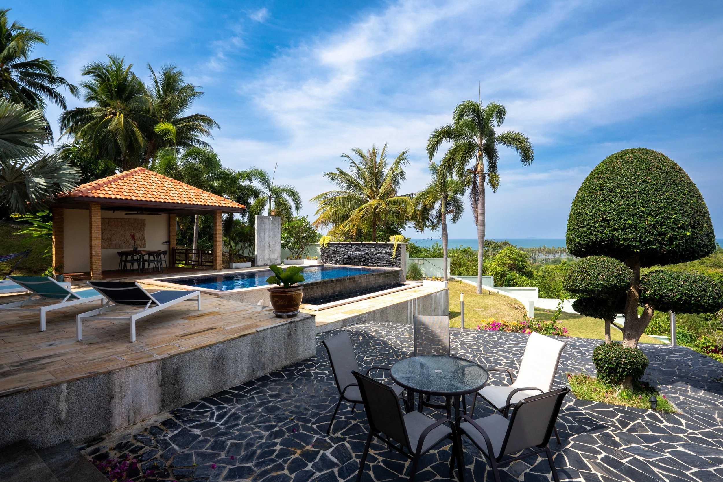 A luxurious outdoor area with a swimming pool, lounge chairs, a shaded gazebo, and manicured trees, overlooking a green landscape and the ocean in the distance.