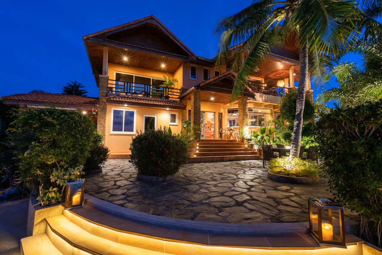 Nighttime view of a well-lit two-story house with a tiled roof, surrounded by lush tropical plants, palm trees, and a stone pathway.