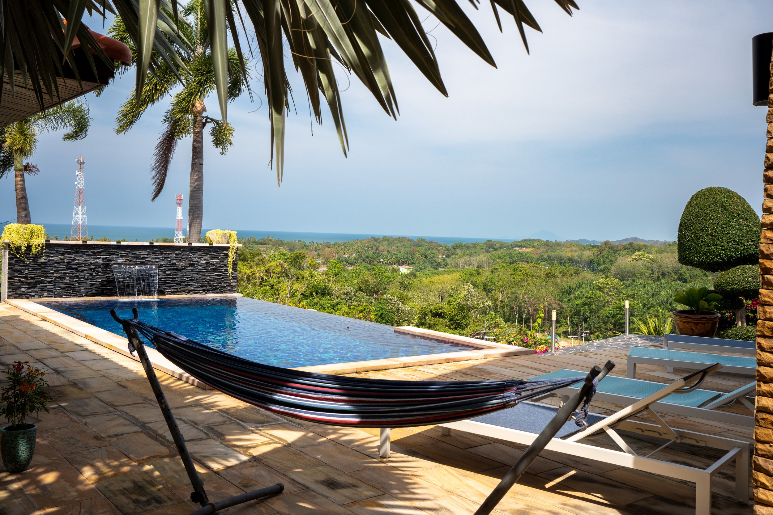 A backyard patio with an infinity pool overlooking a lush green landscape and distant ocean, shaded by palm trees, with lounge chairs and a hammock.