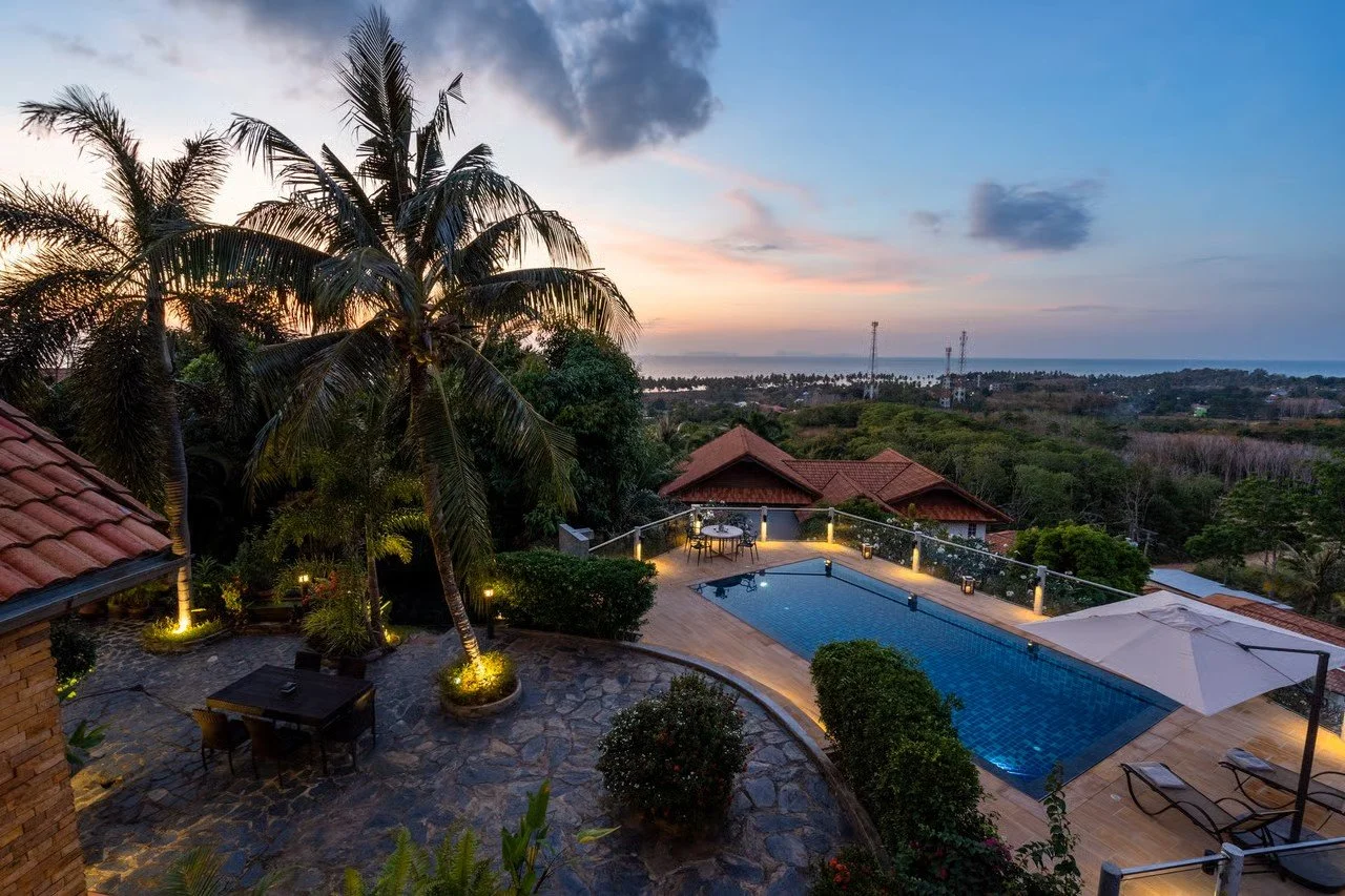 A scenic view of a backyard with a swimming pool, palm trees, outdoor dining area, and sunset sky.