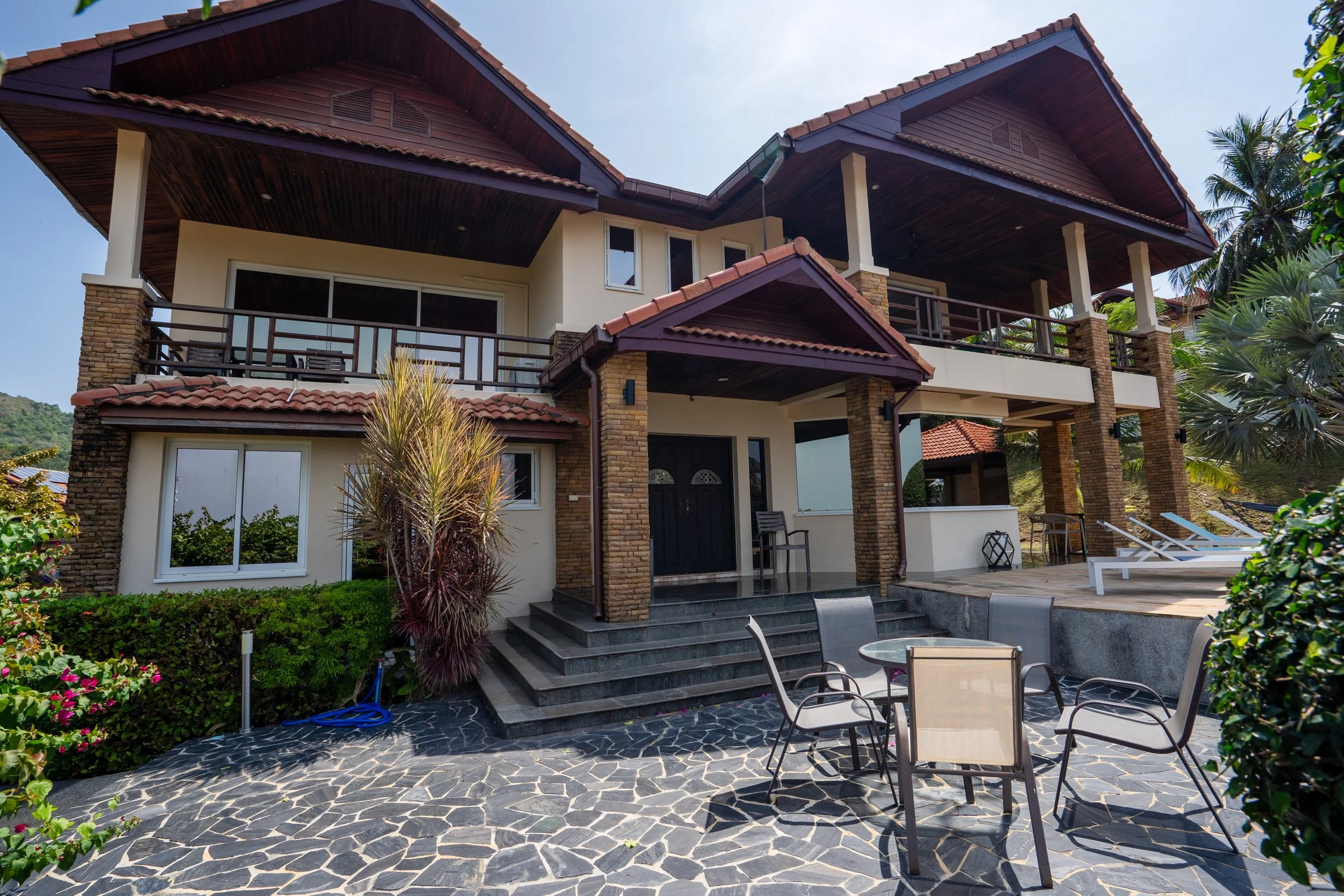 Front view of a two-story house with a stone and stucco exterior, a tiled roof, balcony, and patio area with outdoor furniture, surrounded by greenery.