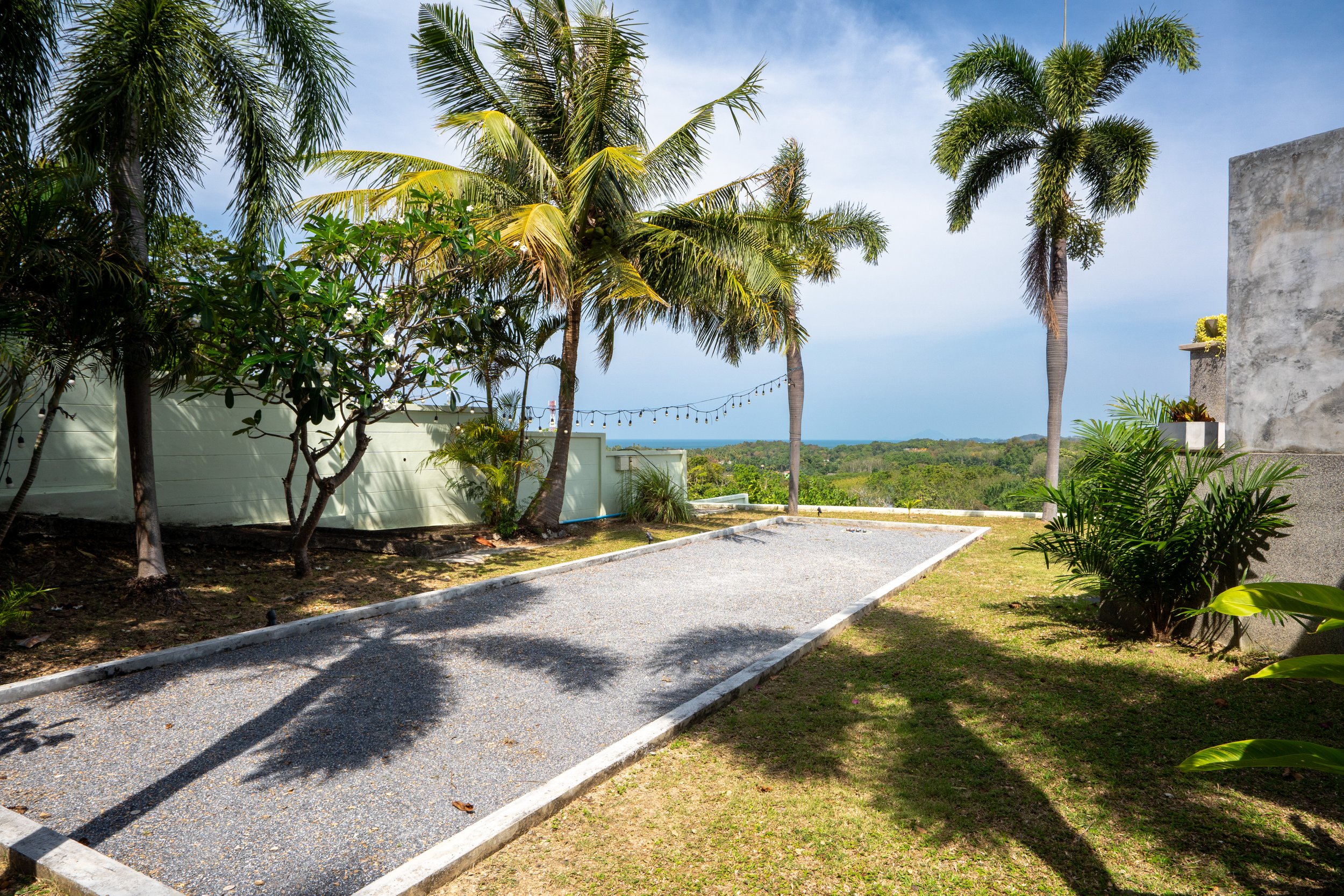 A gravel game court surrounded by tropical plants and palm trees, with string lights hanging across the background and a view of a tree-lined landscape with the ocean in the distance.