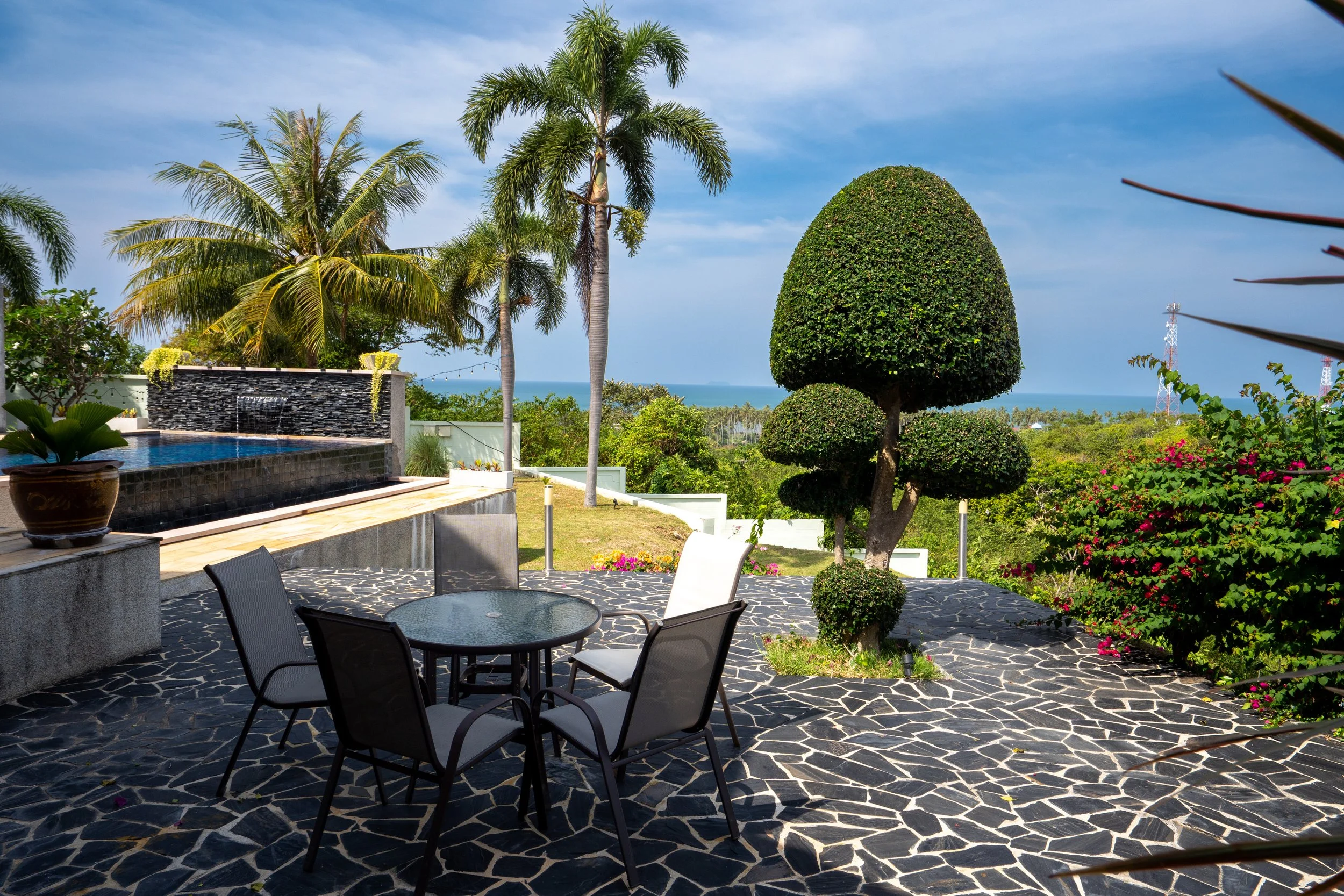 Outdoor patio with black and white stone flooring, surrounded by lush green trees, including palm trees and a topiary, with a view of the ocean in the background, and a small round glass table with four chairs.