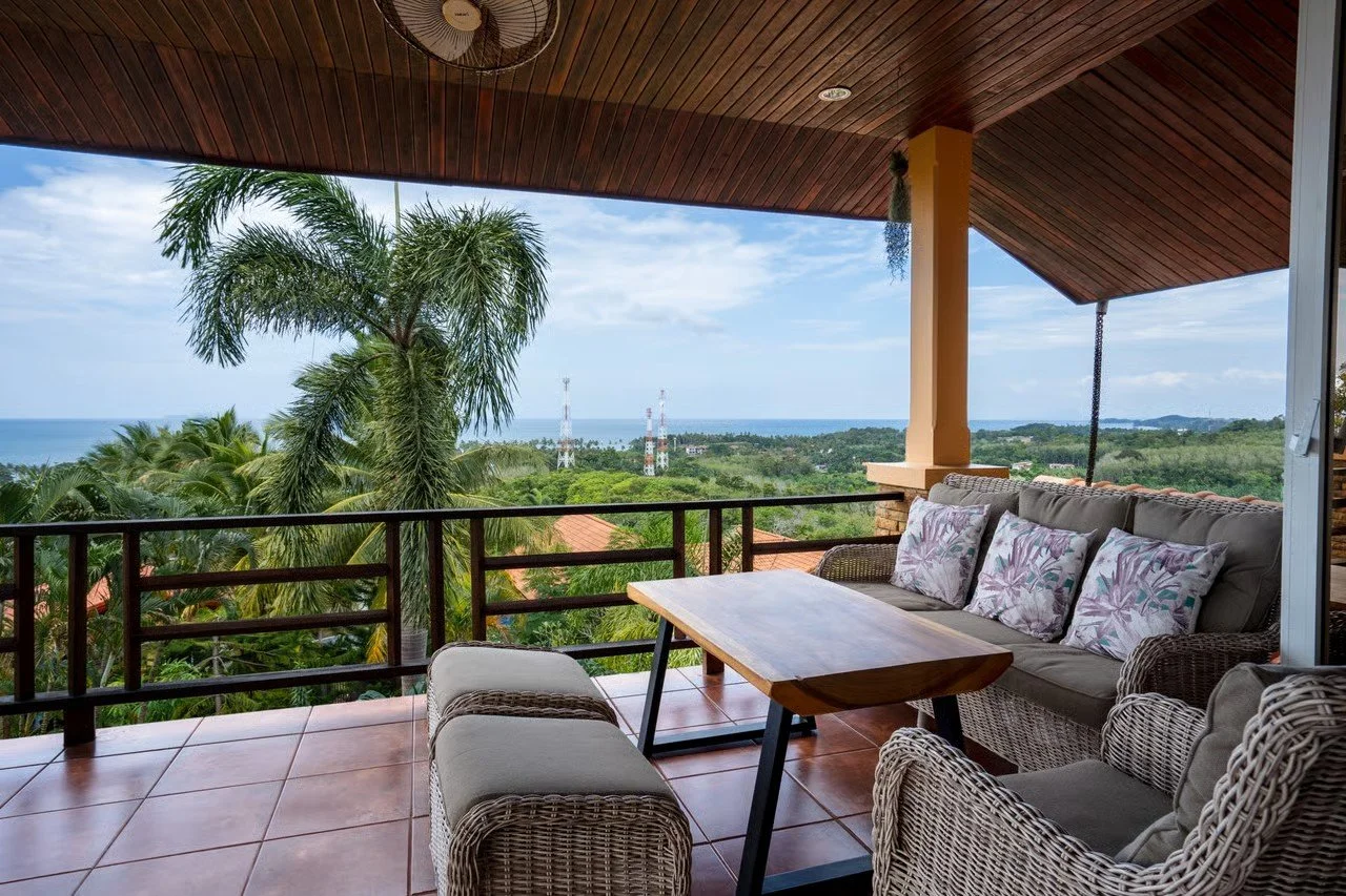 Balcony with wicker chairs and a sofa with floral cushions, overlooking lush green trees, palm trees, and the ocean in the distance under a partly cloudy sky.