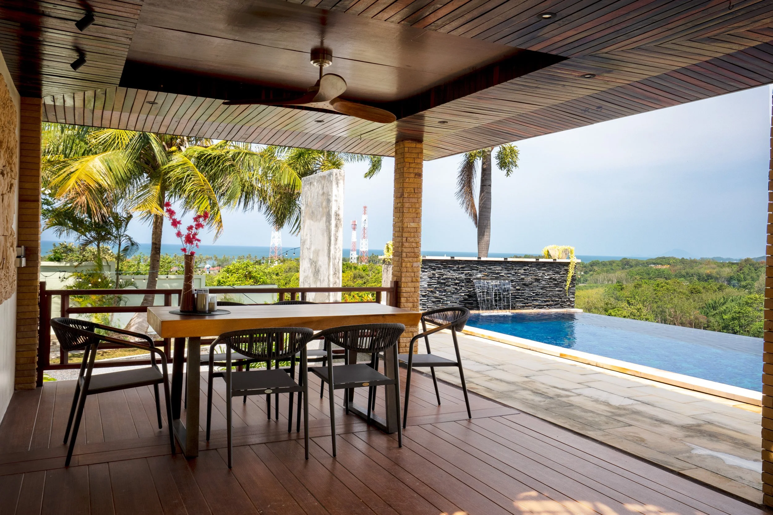 Outdoor patio with a dining table and chairs, overlooking a swimming pool with a waterfall feature, surrounded by tropical trees and a view of the ocean in the distance.