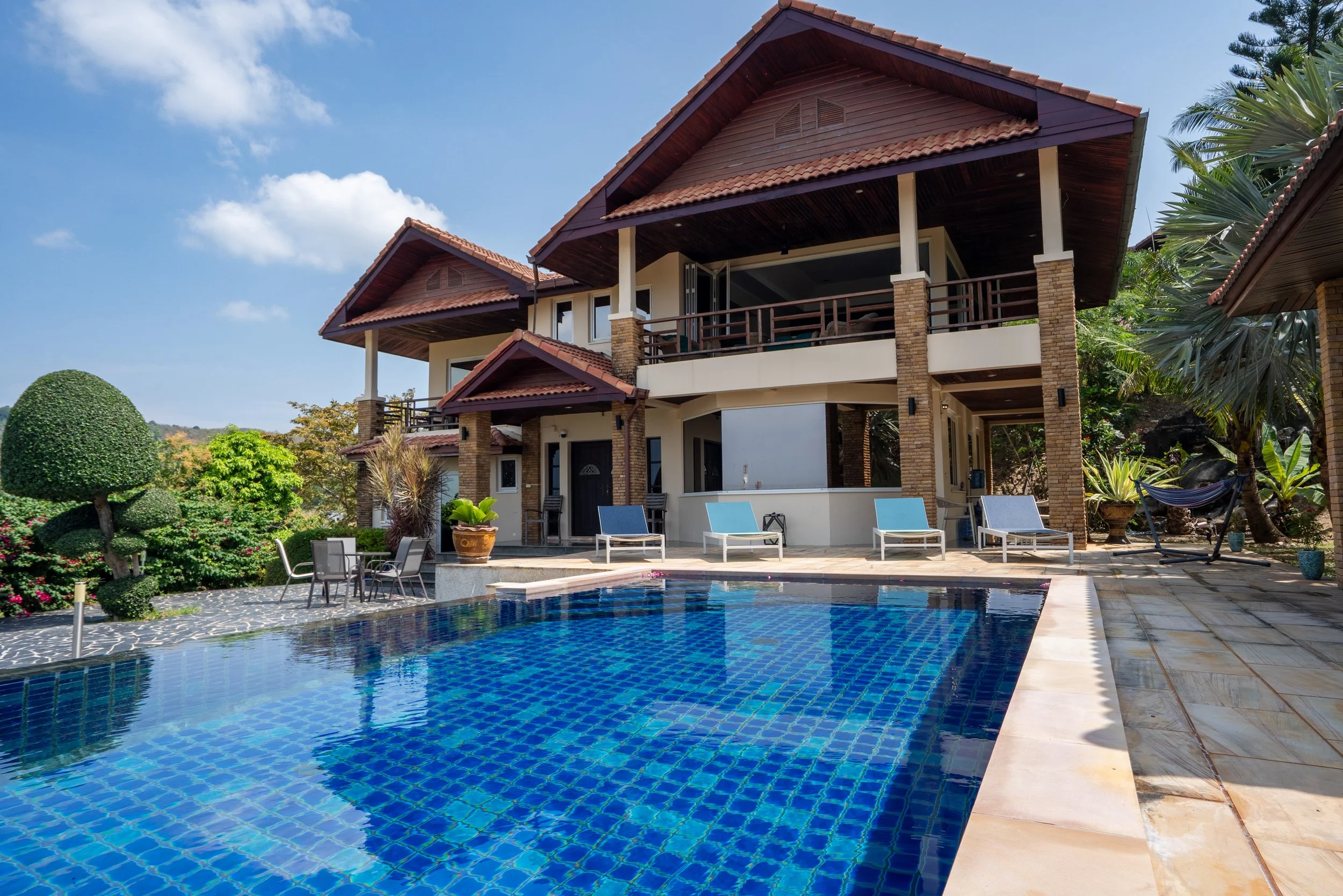 A large two-story house with a swimming pool in the foreground, surrounded by lush greenery and outdoor furniture, under a partly cloudy sky.