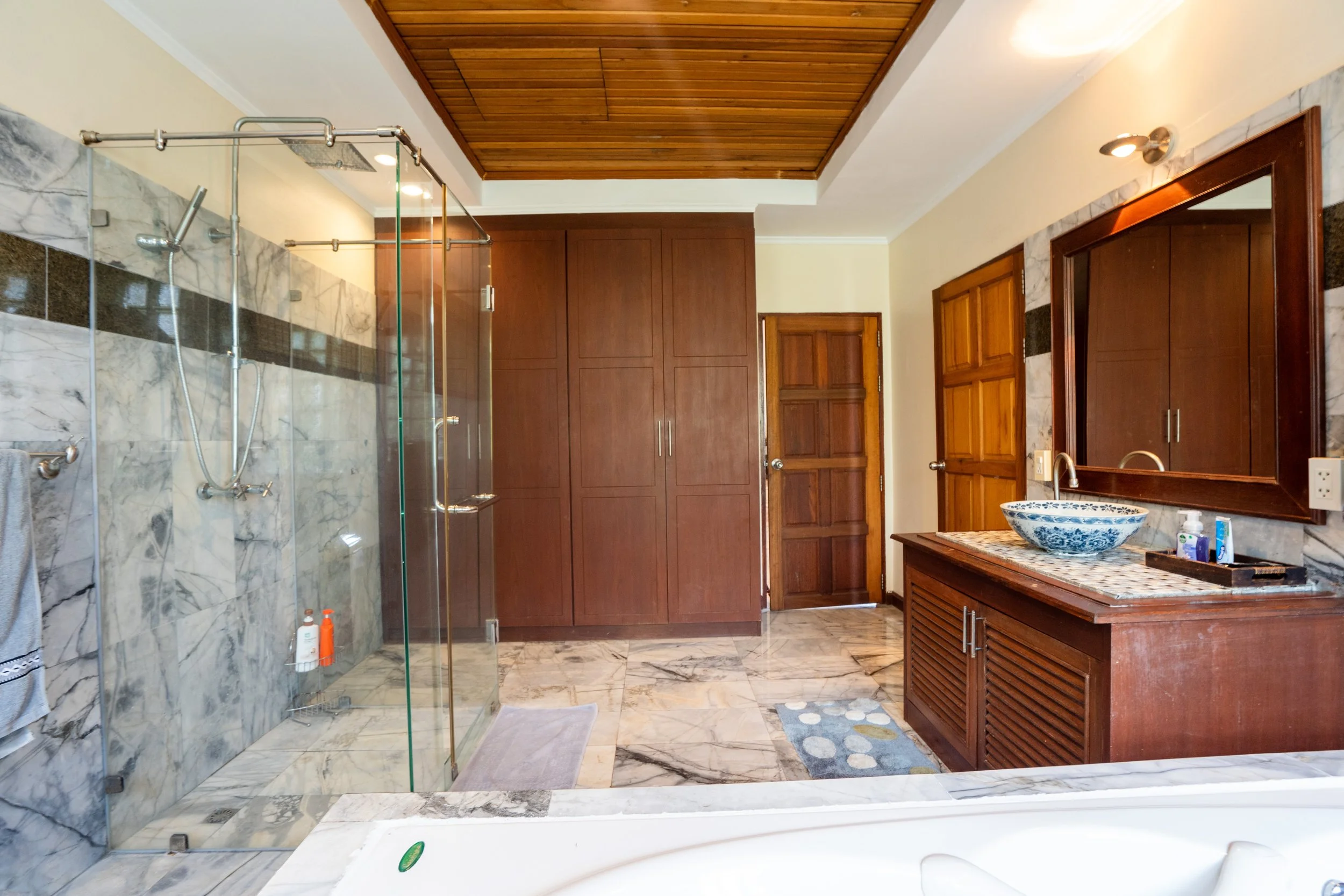 A spacious bathroom featuring a glass-enclosed shower with marble tiles, wooden cabinetry, a decorative vessel sink, and a large mirror. The room has a wooden ceiling and a closet area with double wooden doors.