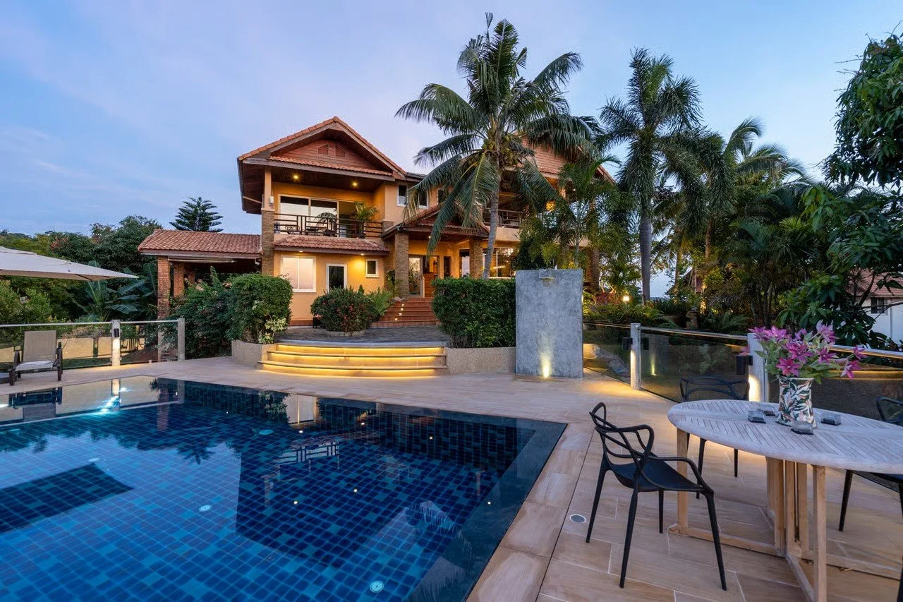 Luxury house with a swimming pool, outdoor seating area, and tropical landscaping at dusk.