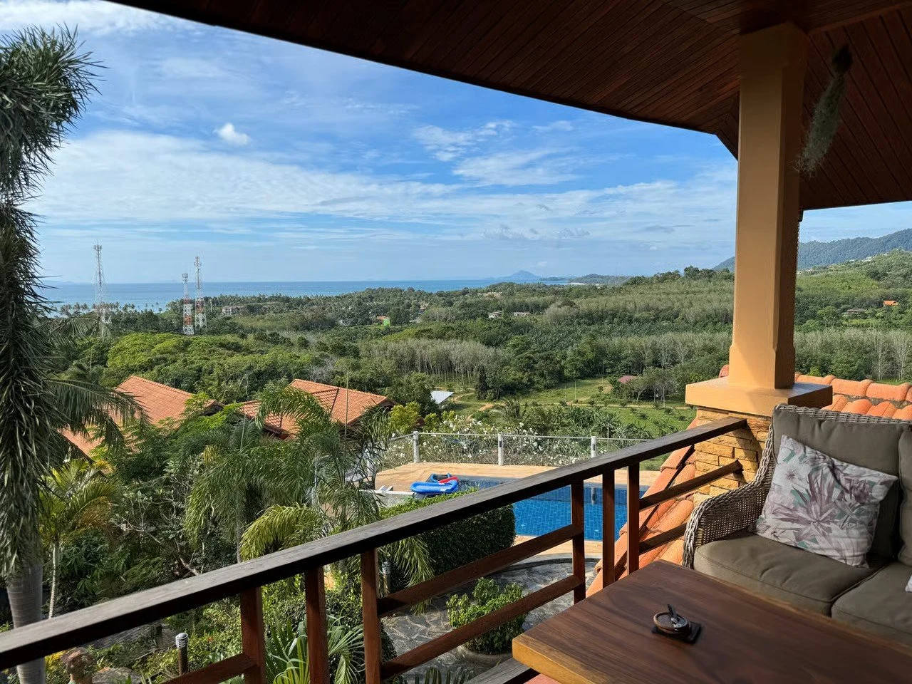 Balcony view overlooking a lush landscape with trees, rooftops, and hills, with the ocean and blue sky in the distance. Outdoor furniture and a swimming pool are visible in the foreground.