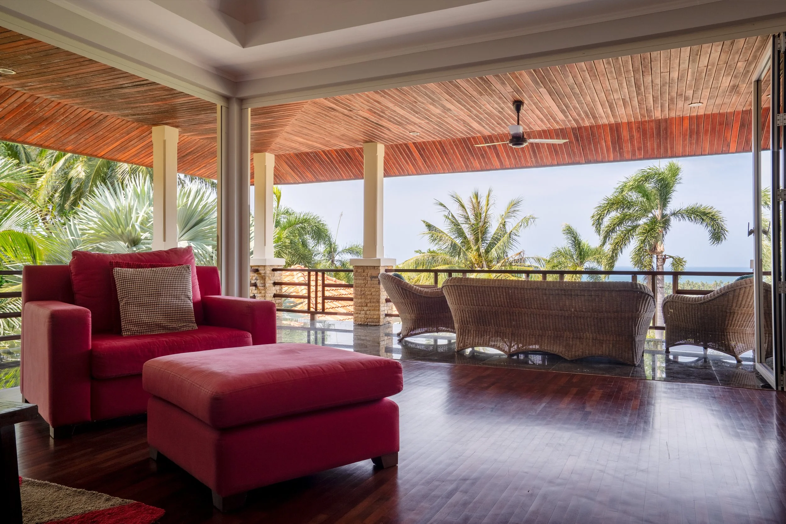 Cozy outdoor balcony with red armchair and ottoman, wicker furniture, tropical palm trees, wooden ceiling, and ocean view in the background.