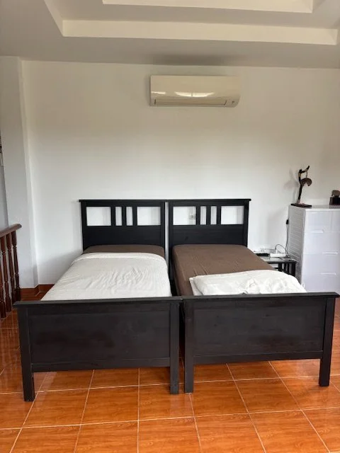 A room with two twin beds with black wooden headboards, a white air conditioning unit on the wall, a small white dresser, and a wooden side table with a decorative sculpture.