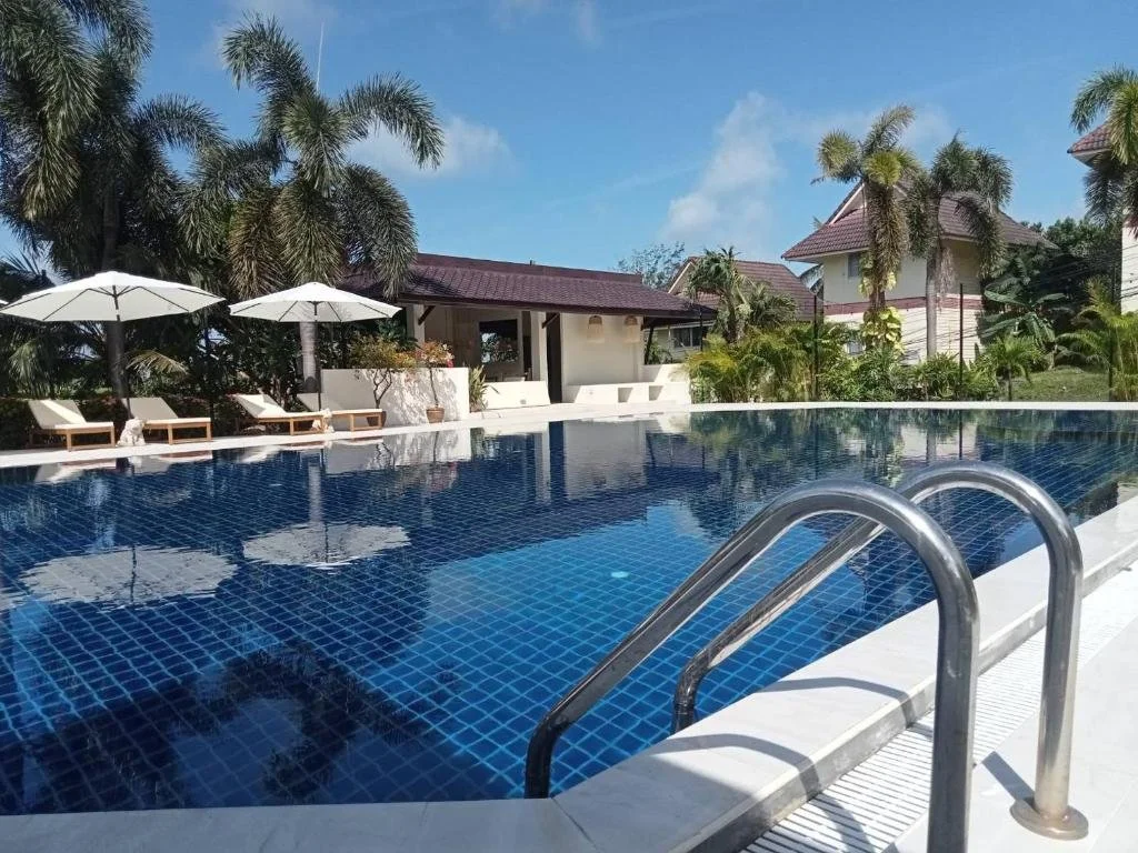 A swimming pool with blue tiles, surrounded by lounge chairs with umbrellas, tropical palm trees, and buildings in the background, under a partly cloudy sky.
