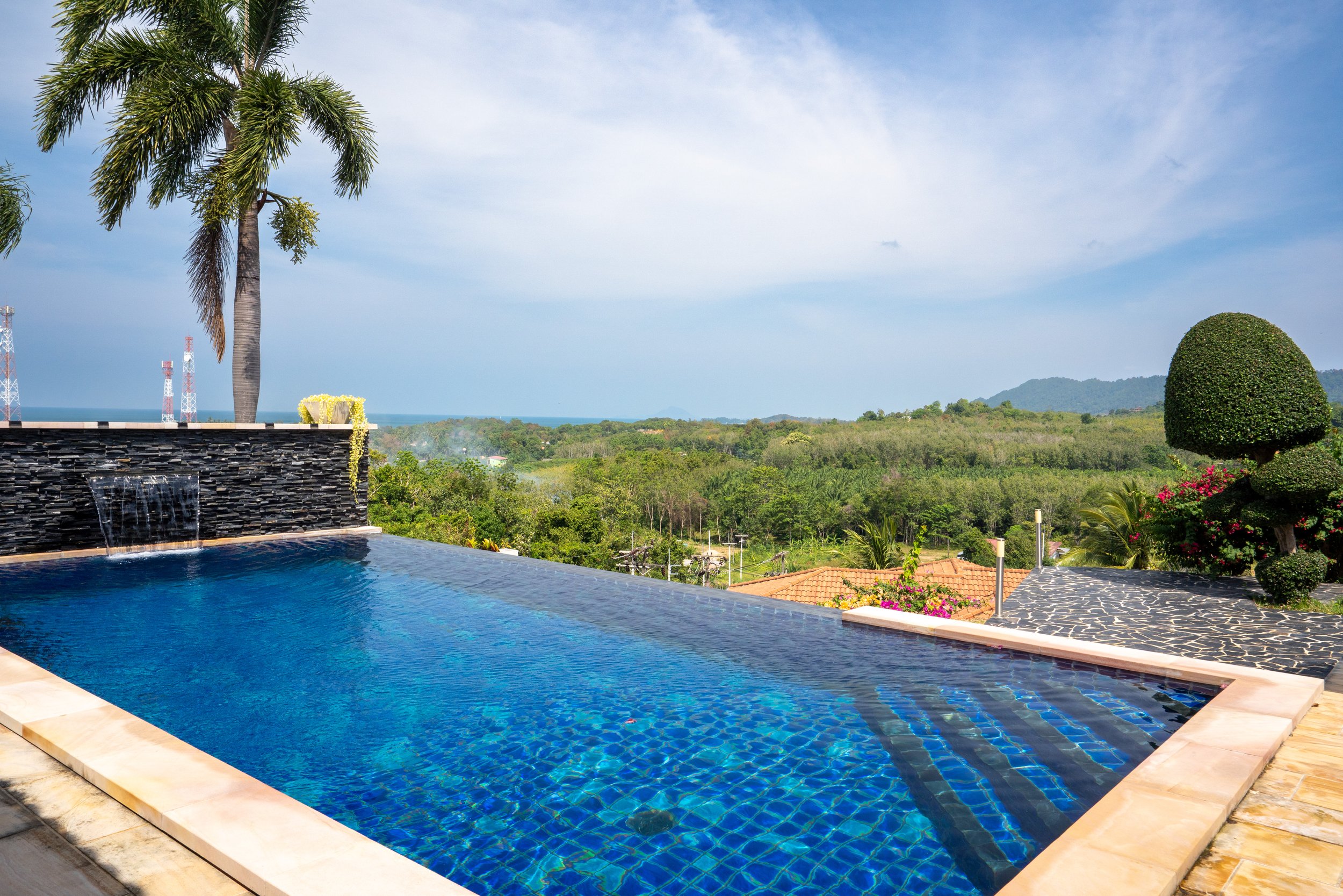 Swimming pool with waterfall feature, surrounded by tropical plants, palm trees, and a scenic view of distant green hills and blue sky.